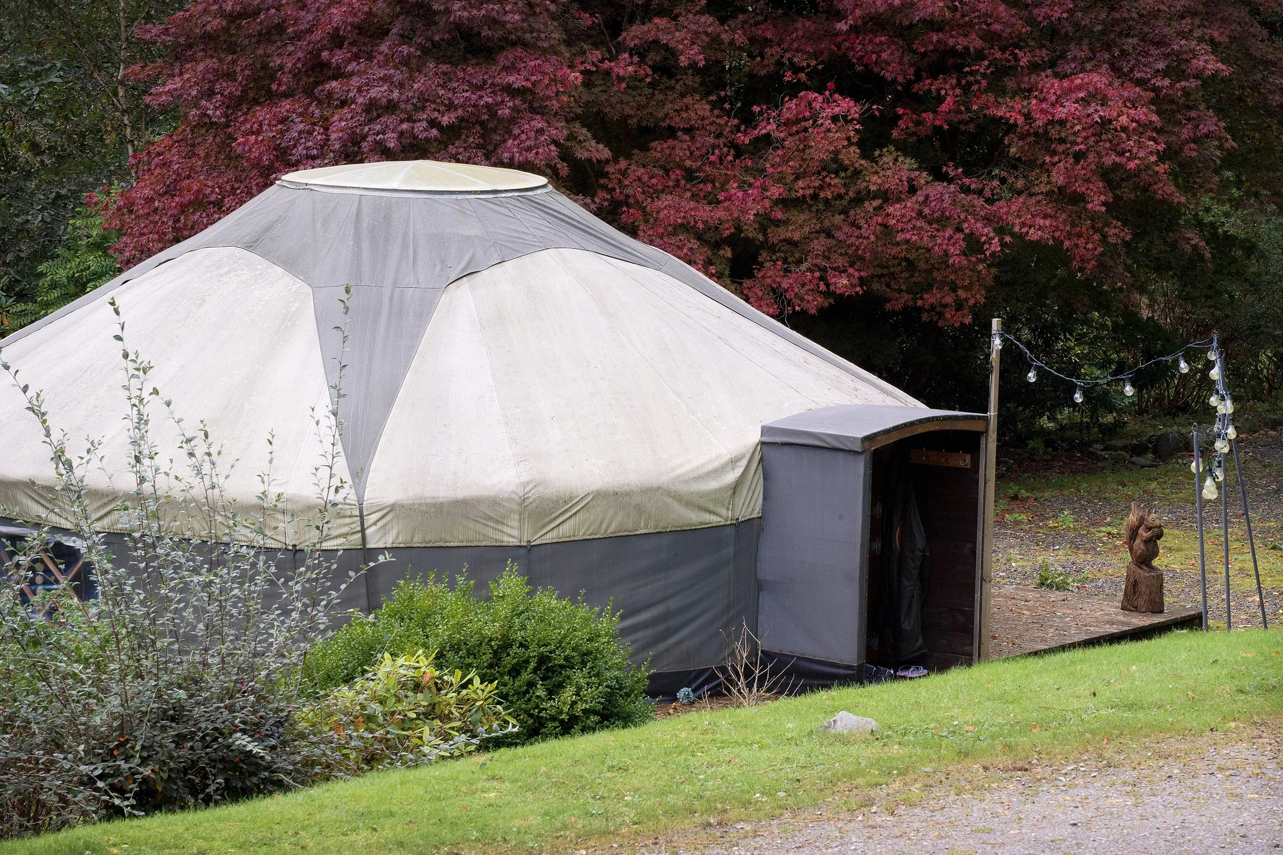 A large circular tent or yurt with a beige and gray exterior, set outdoors on a grassy area with surrounding bushes and trees. There are string lights on poles and a wooden sculpture nearby.