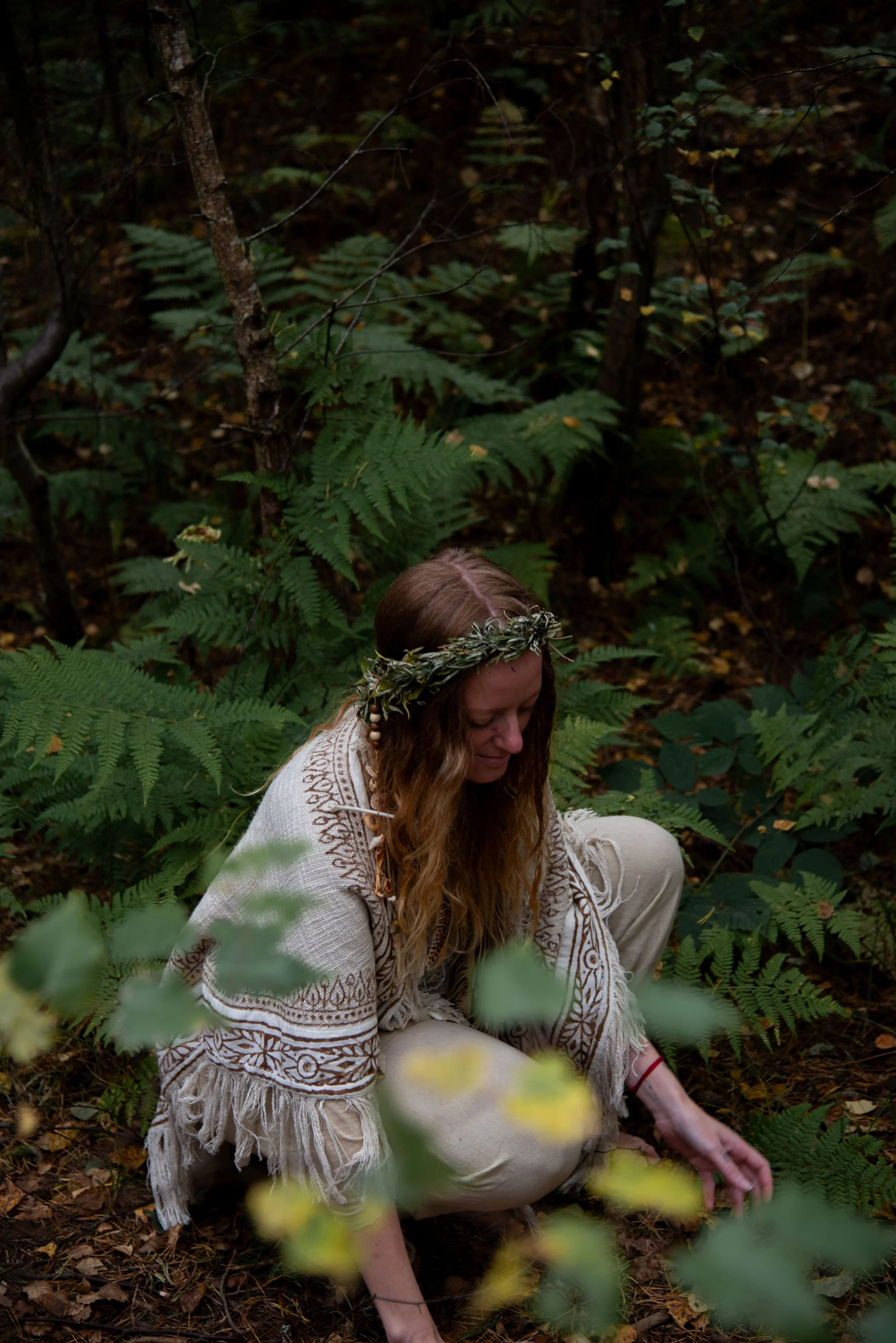 A woman with long, wavy brown hair wearing a white, embroidered shawl and a laurel wreath on her head, crouching in a forest surrounded by green ferns and trees.