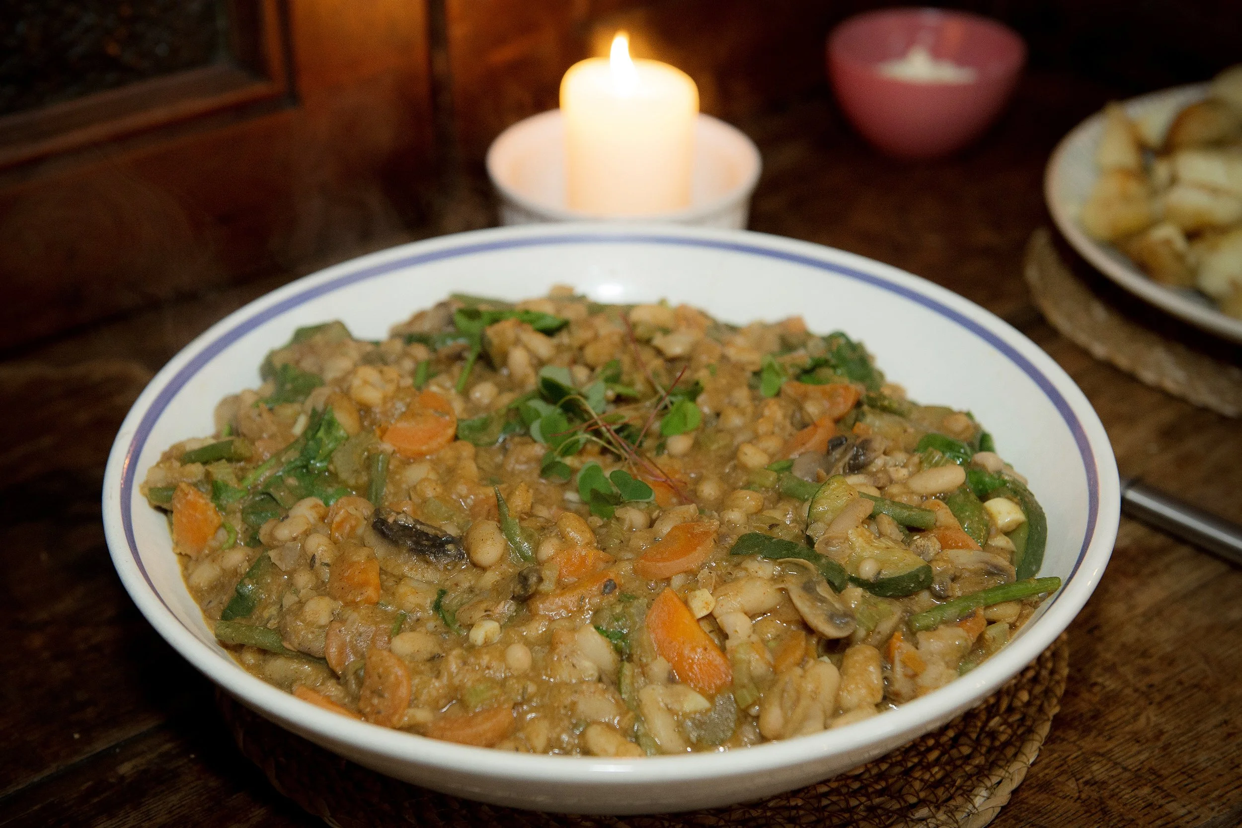 A bowl of hearty vegetable stew with carrots, green beans, and other vegetables on a wooden table, lit by a candle in the background.
