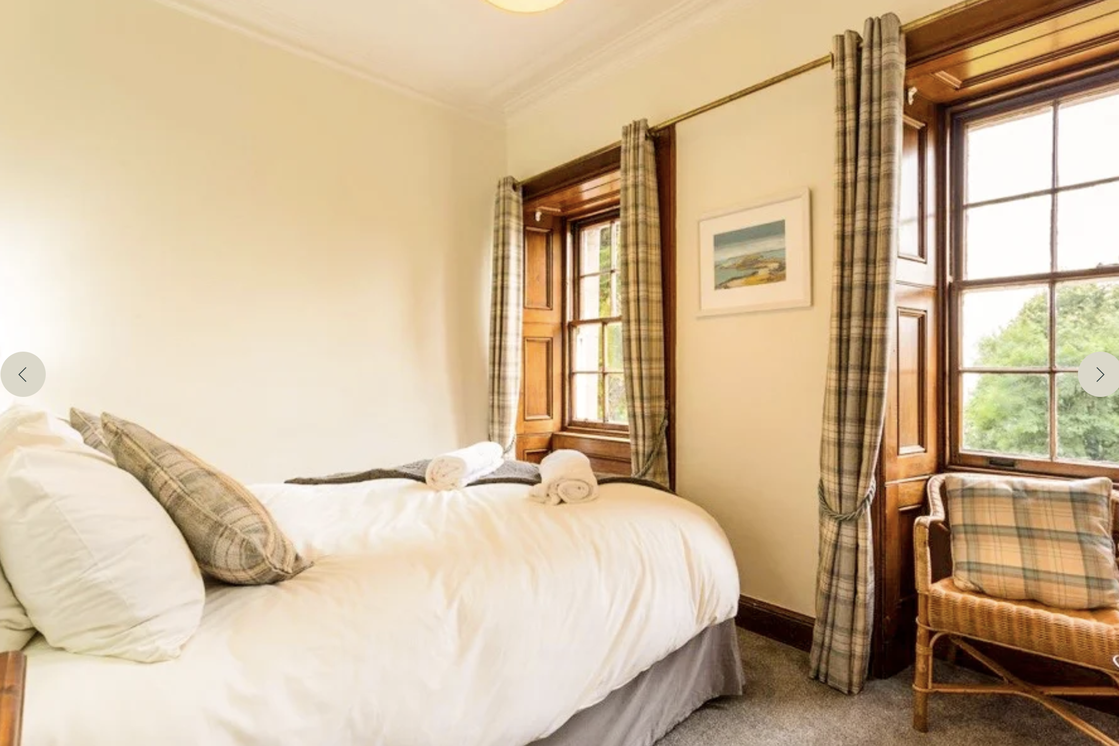 A cozy bedroom with a bed topped with white linens, plaid pillows, and rolled towels. Natural light streams through two wooden-framed windows dressed with plaid curtains, and a framed landscape painting hangs on the beige wall. There is a wooden chair with a plaid cushion in the corner.
