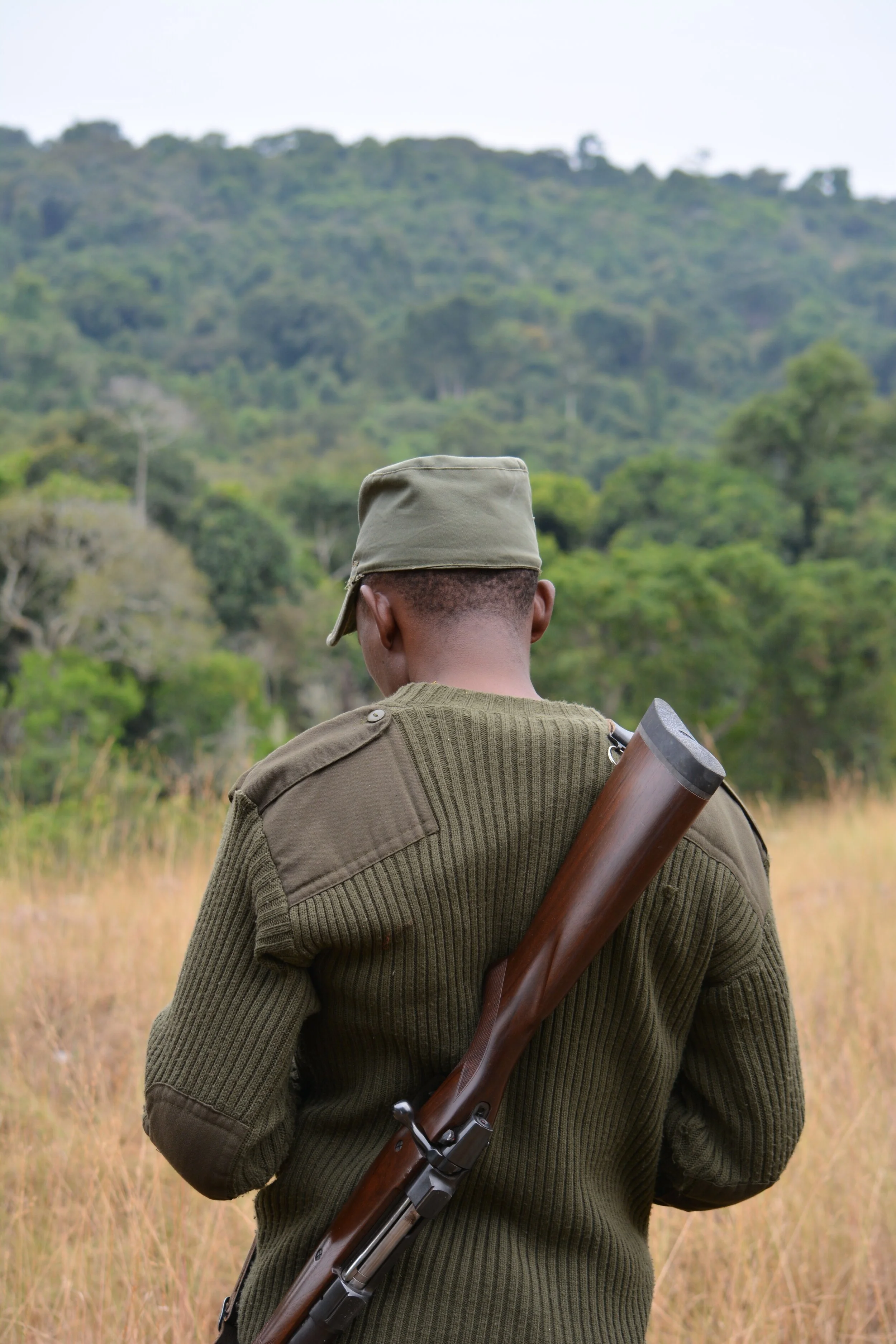 Person in military uniform holding a rifle, standing in a grassy field with a forested hill in the background.