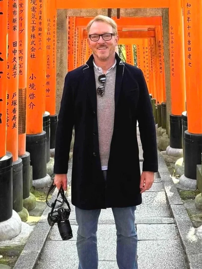 A man standing on a stone pathway in front of orange torii gates at a Japanese shrine. He is smiling, wearing glasses, a gray sweater, a dark coat, and holding a camera in his right hand.