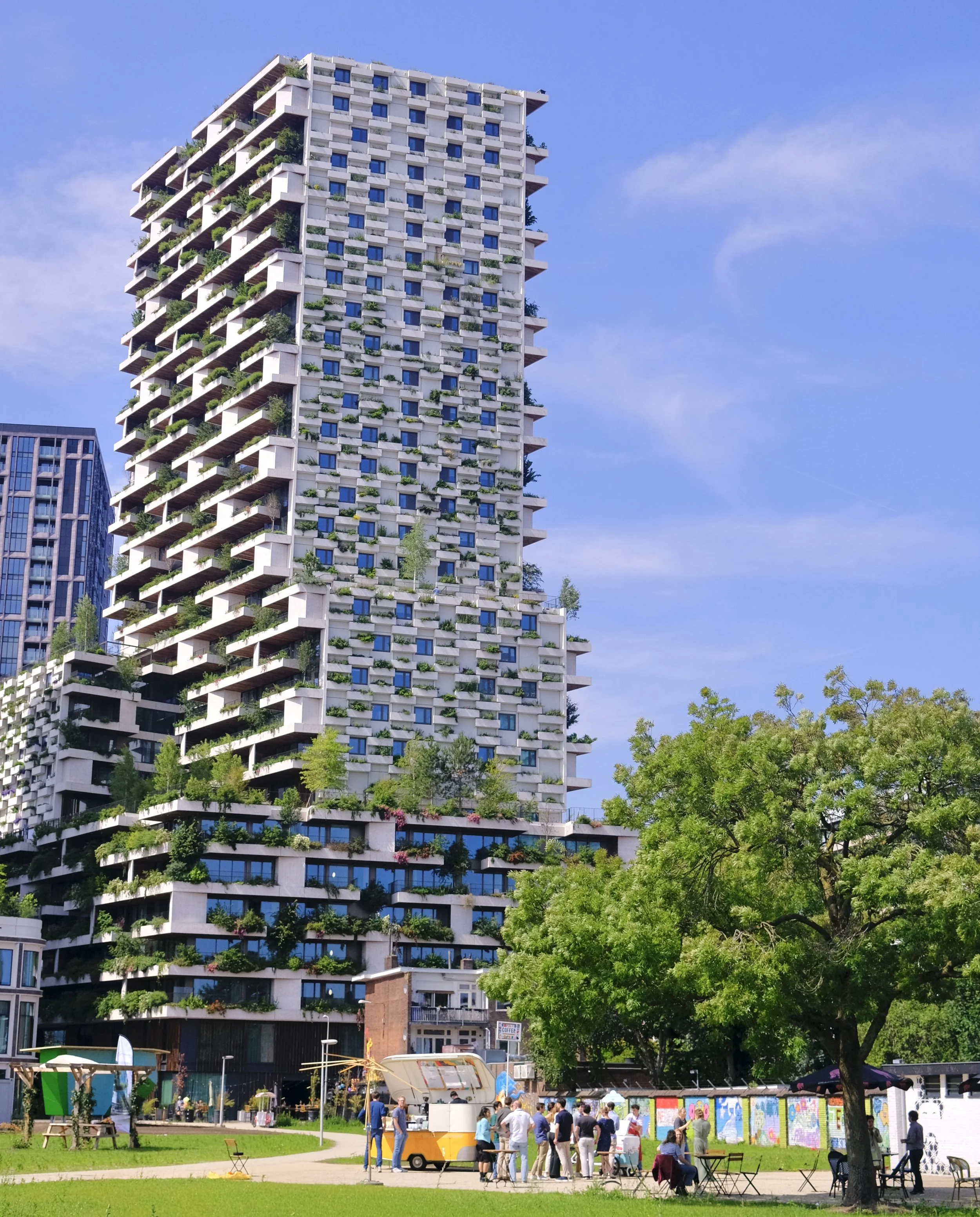People gathered around a food truck and outdoor tables in a park with a tall modern apartment building in the background, blue sky, and lush green trees.