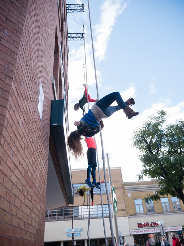 Four performers are wrapped in canvas-covered ropes, hanging from the side of a building, about a story-high. Three are white, one is queer, and one is a Colombian amputee. Some are upside down and some are upright. They wear bright  colours.