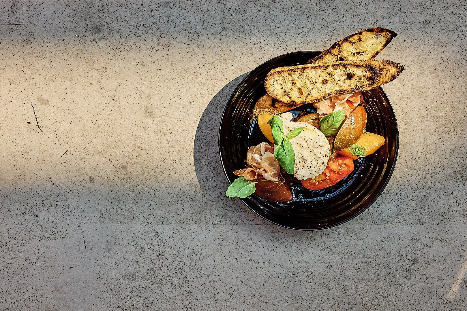 A bowl of salad with tomatoes, basil, and slices of toasted bread on a gray surface.