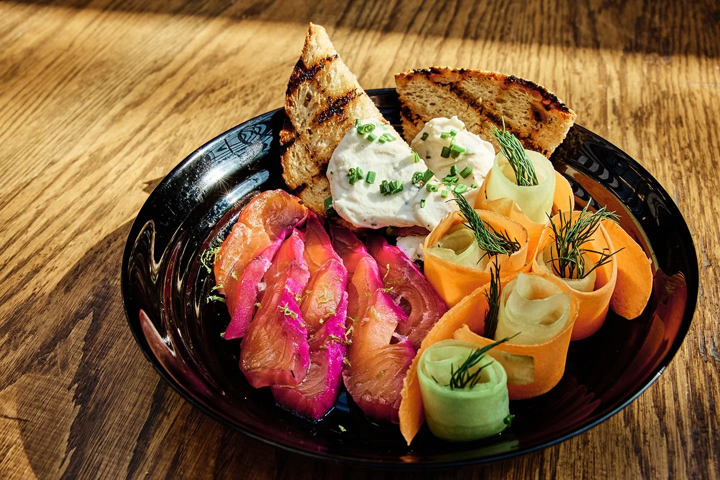 A black plate with smoked salmon, dill, cream cheese, grilled bread slices, and vegetable curls on a wooden table.