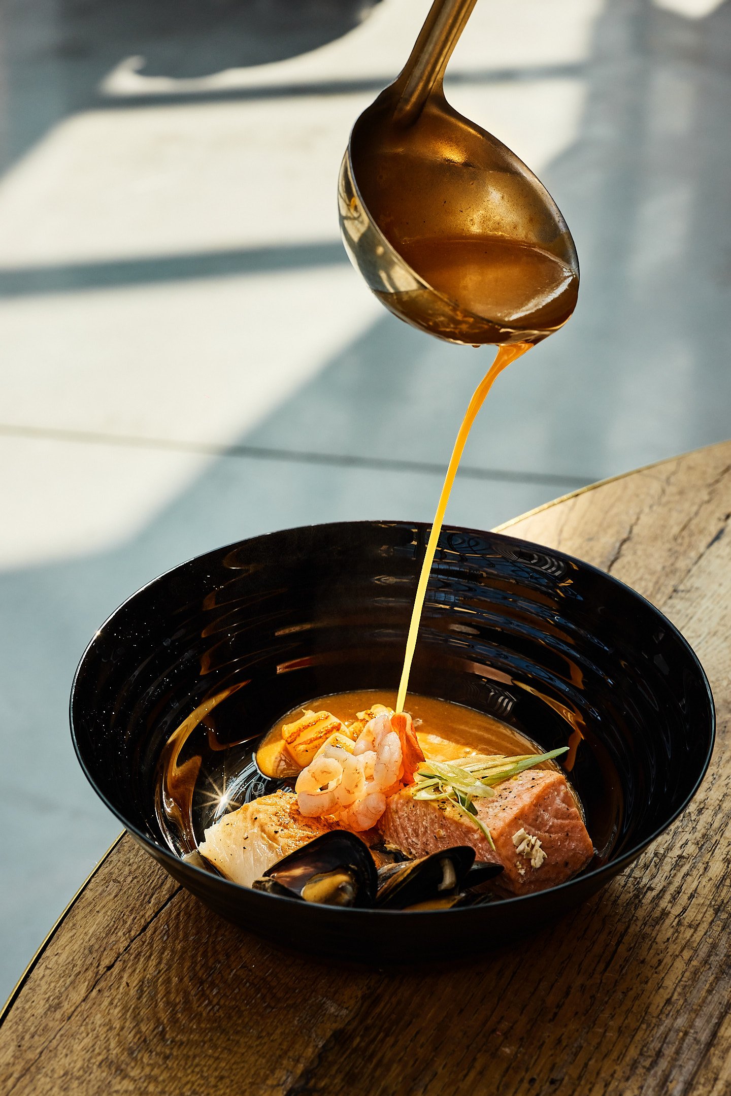 A black bowl containing assorted seafood, with gravy being poured from a spoon over it.