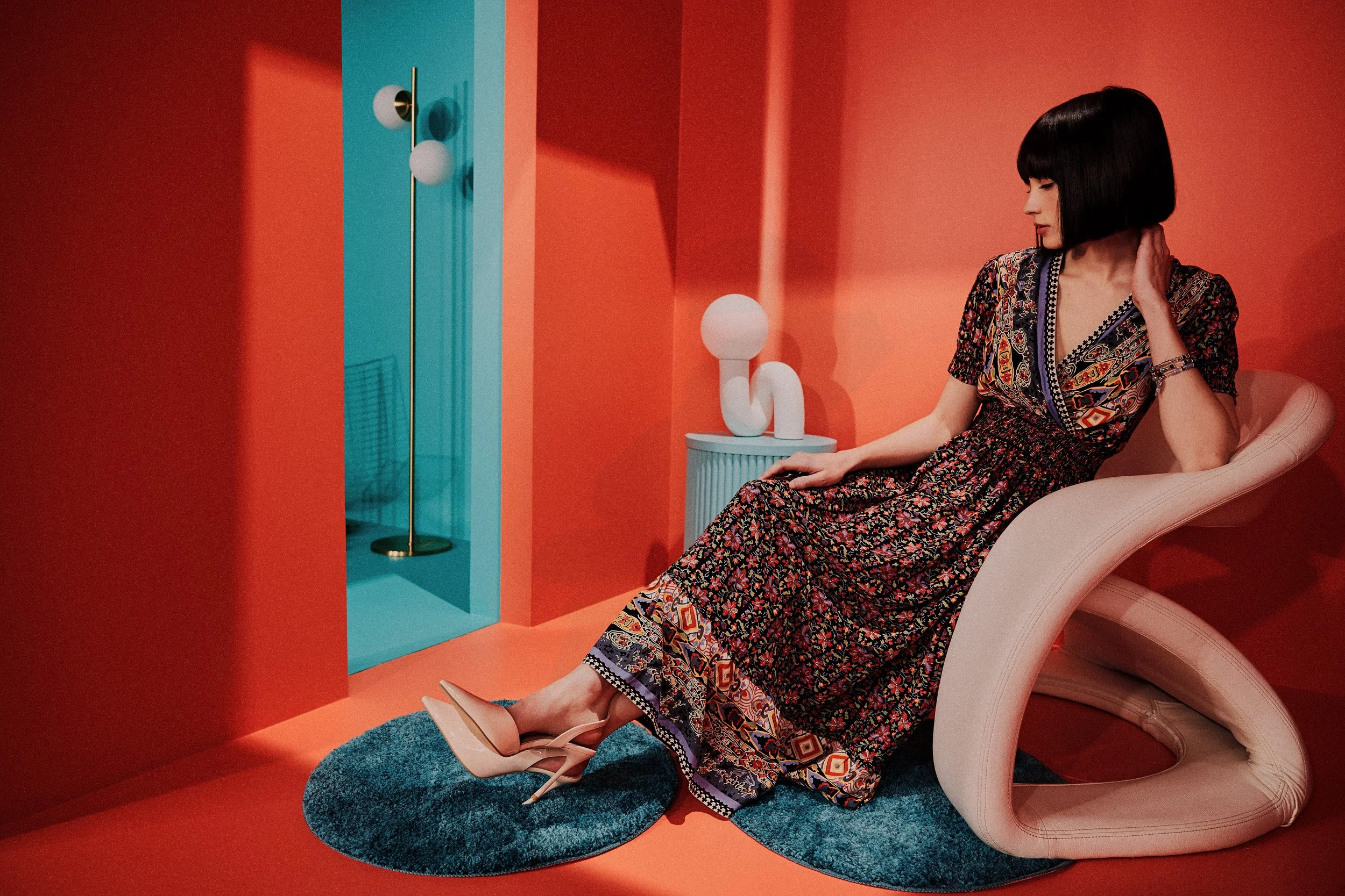 A woman with a black bob haircut sitting on a modern beige lounge chair in a colorful room with orange walls. She is wearing a long floral dress and beige high heels, with her legs extended onto a circular teal rug. There is a small round side table with a white abstract sculpture on it and a tall gold floor lamp with three white globes in the background.