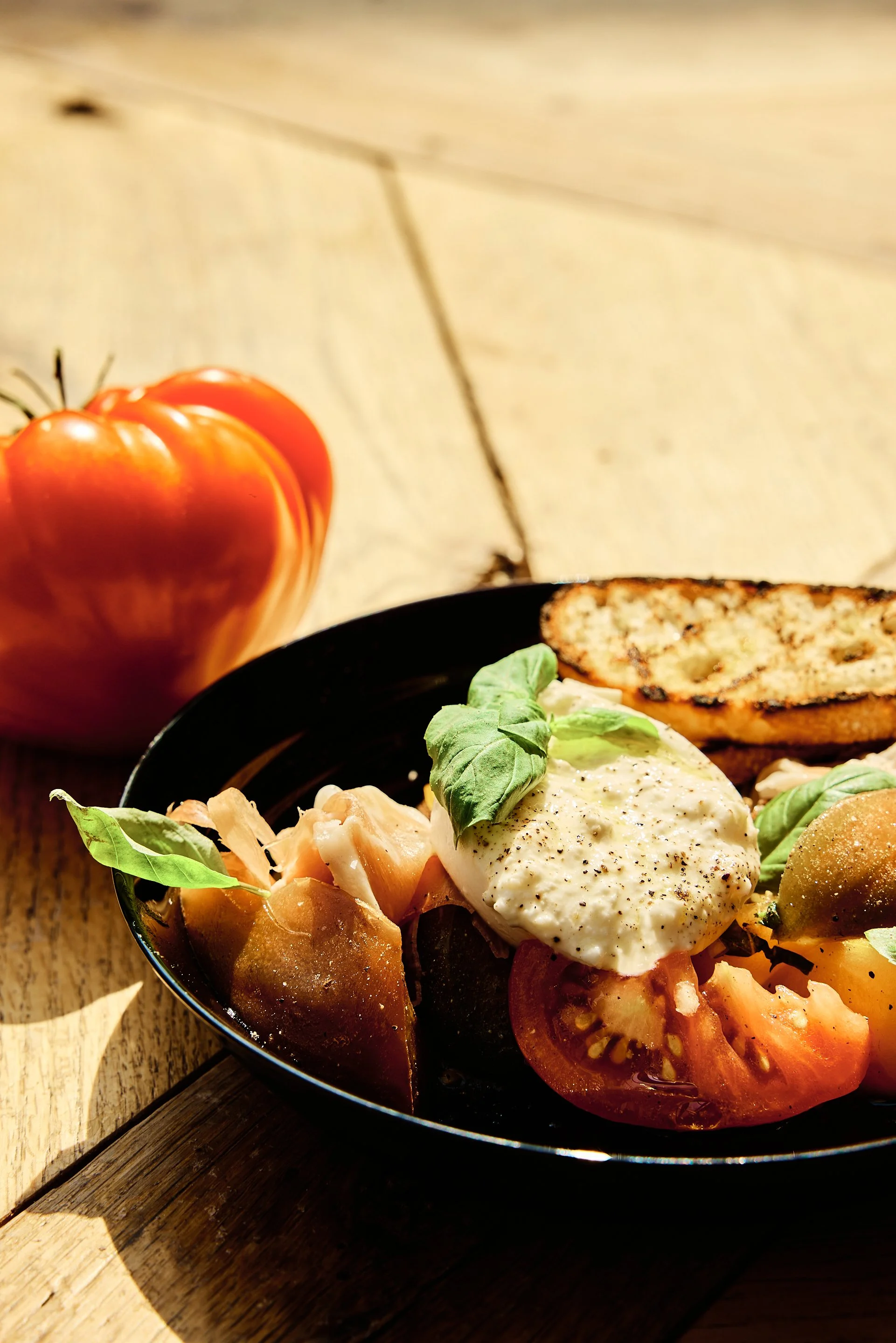 A bowl of fresh tomato salad topped with a poached egg and basil leaves, with toasted bread on the side, set on a wooden table outdoors with sunlight.