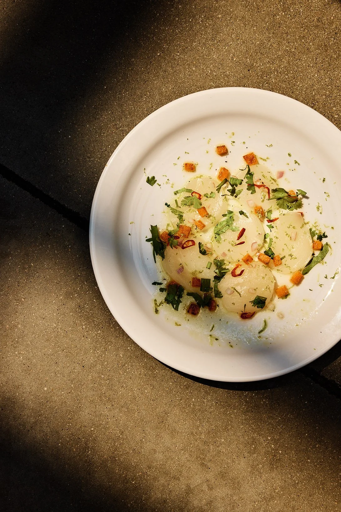 A white plate with peeled potatoes garnished with chopped herbs, small crispy croutons, and red pepper flakes, placed on a dark surface.