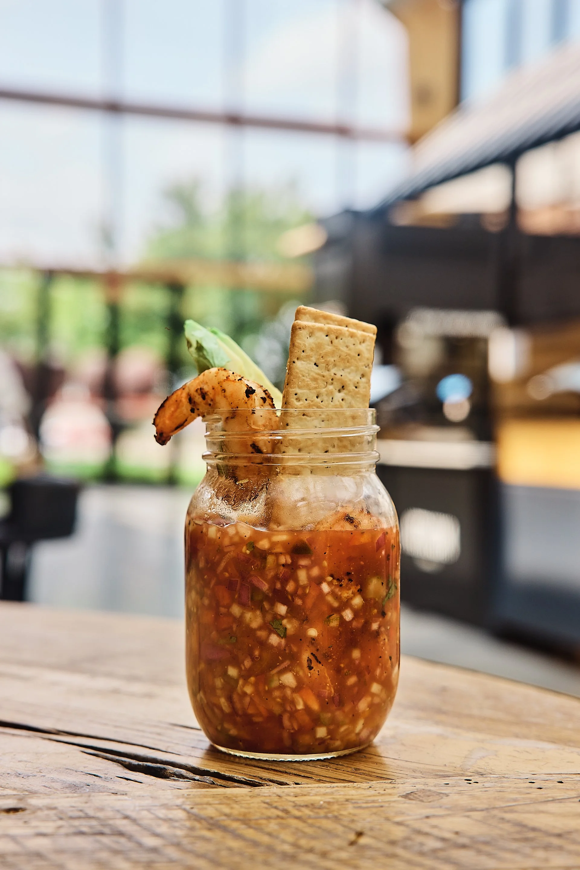 A mason jar filled with chopped vegetables and a tomato-based broth, garnished with a fried plantain, a slice of avocado, and a rectangular crisp wafer, on a wooden table with a blurred indoor background.