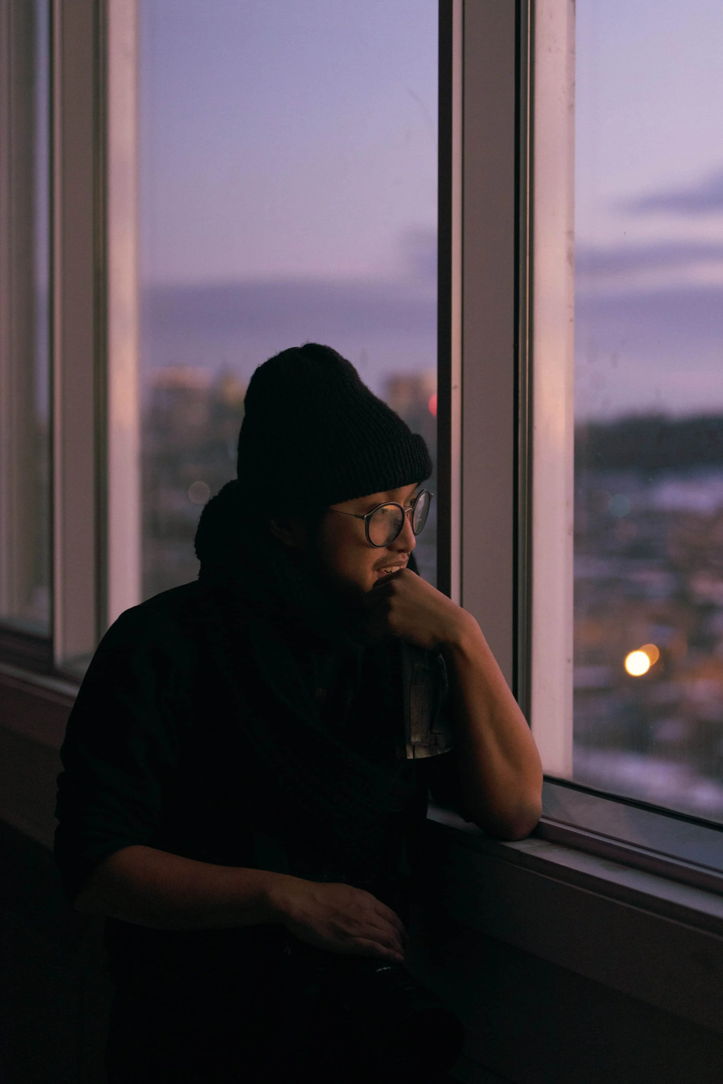 Person wearing a beanie and glasses looks thoughtfully out a window at sunset, with a cityscape blurred in the background.