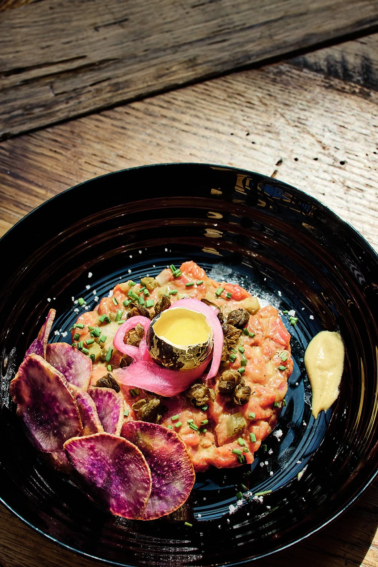Salmon tartare garnished with chopped chives, capers, pickled red onion, a halved quail egg, purple potato chips, and a dollop of mustard, served on a black plate.