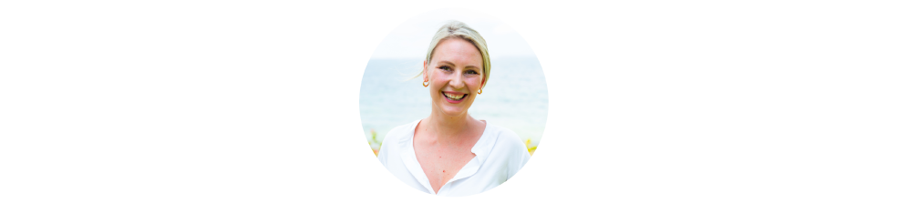 A smiling woman with blonde hair wearing a white top, standing outdoors near a body of water.