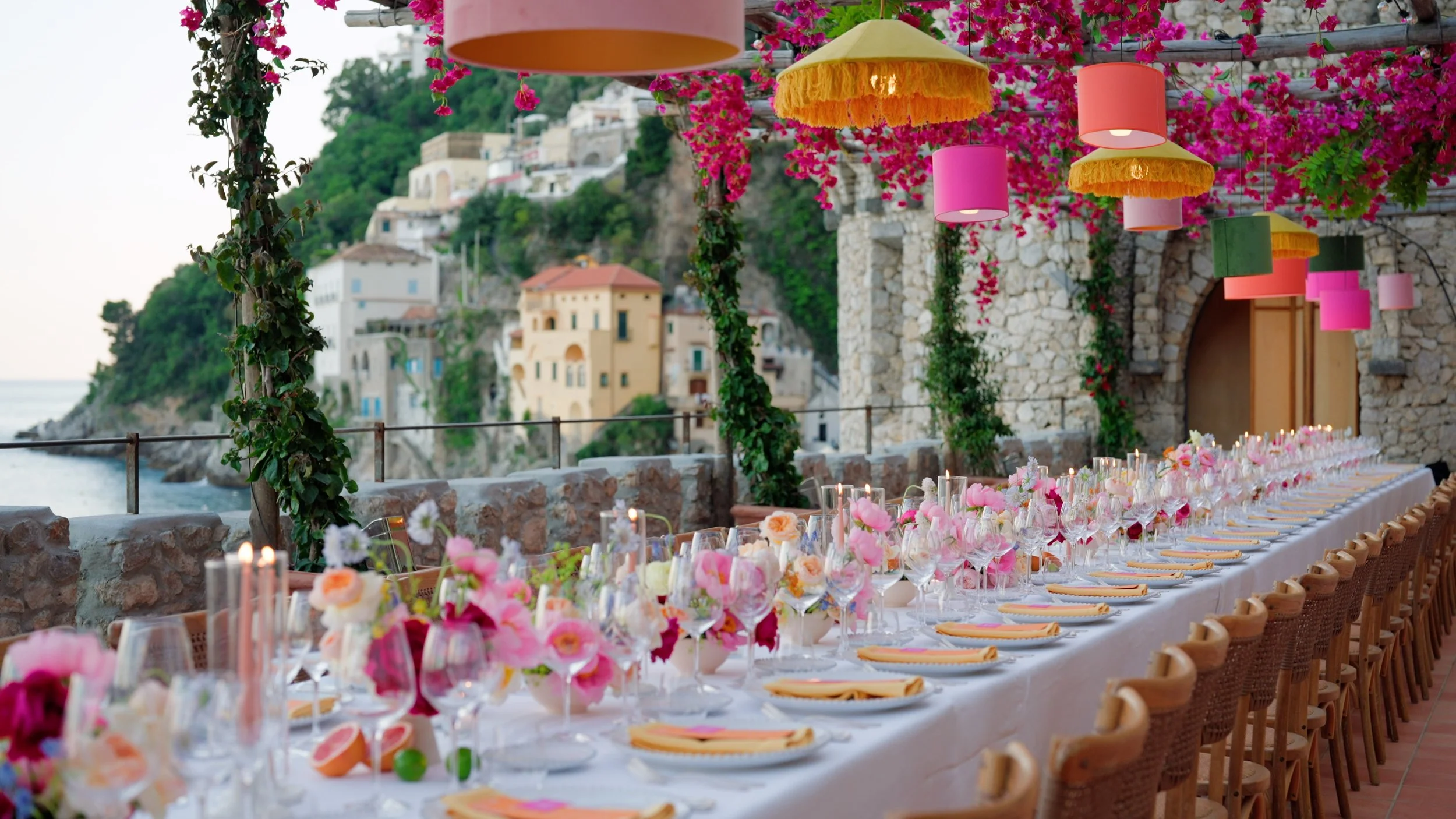 A long outdoor table decorated with pink and orange flowers, candles, and tableware, set for a celebration on a terrace overlooking a Mediterranean coastal town with colorful buildings.