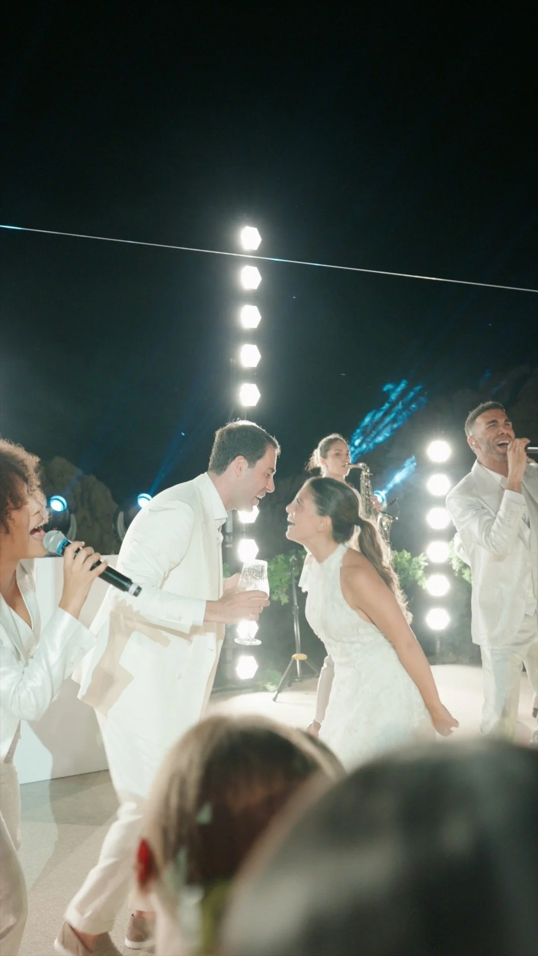 Wedding celebration with people singing, dancing, and playing instruments at night, illuminated by bright lights.