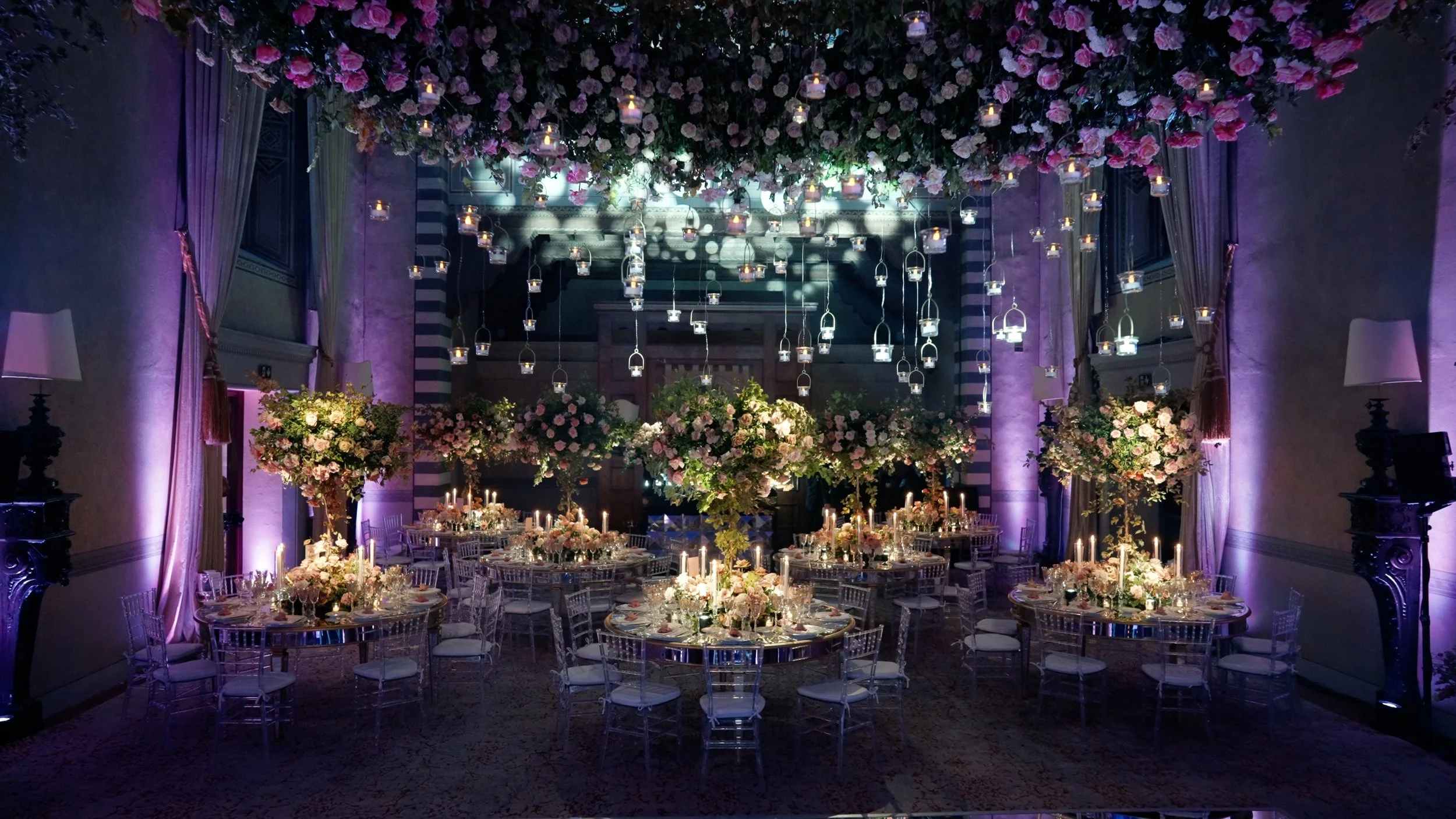 Elegant banquet hall decorated with hanging candles and floral arrangements, featuring round tables with lit candles and chairs covered in white fabric, purple uplighting, and floral topiary centerpieces.