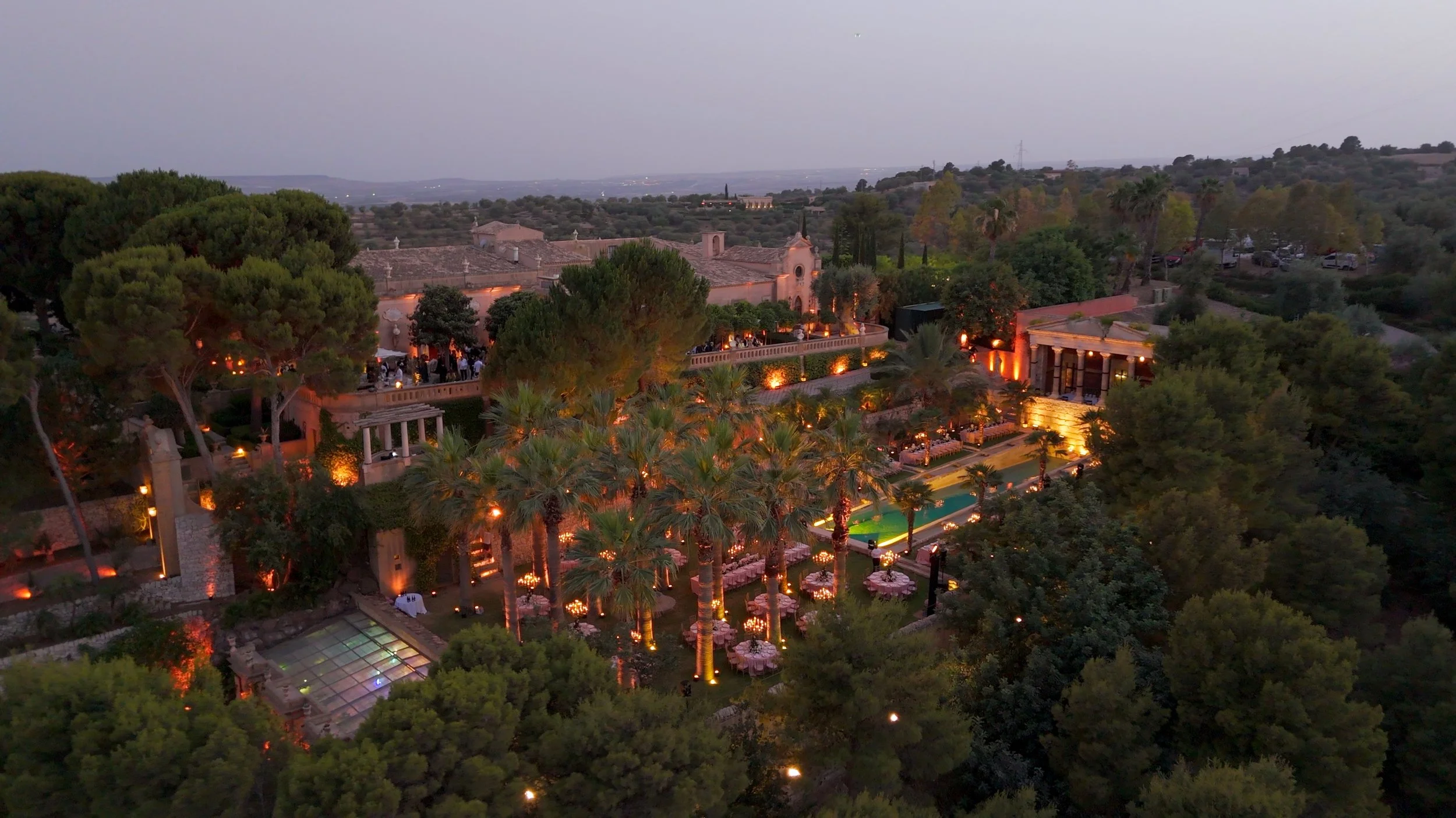 Aerial view of an outdoor event at a luxurious estate with trees, a pool, tables with umbrellas, and warm lighting at dusk.