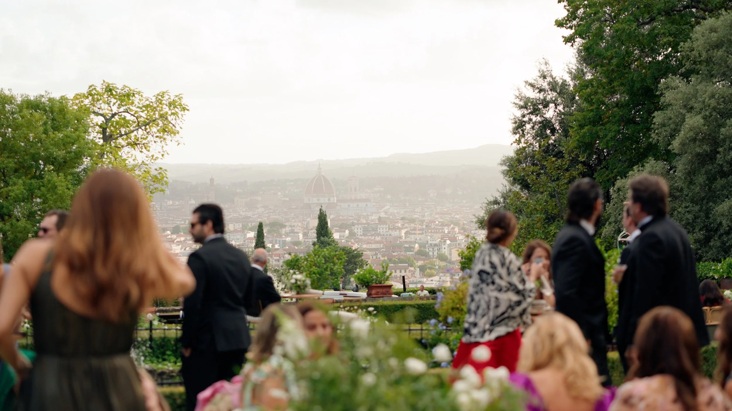 People socializing at an outdoor event with a view of Florence, Italy, including the Florence Cathedral's dome, in the distance.