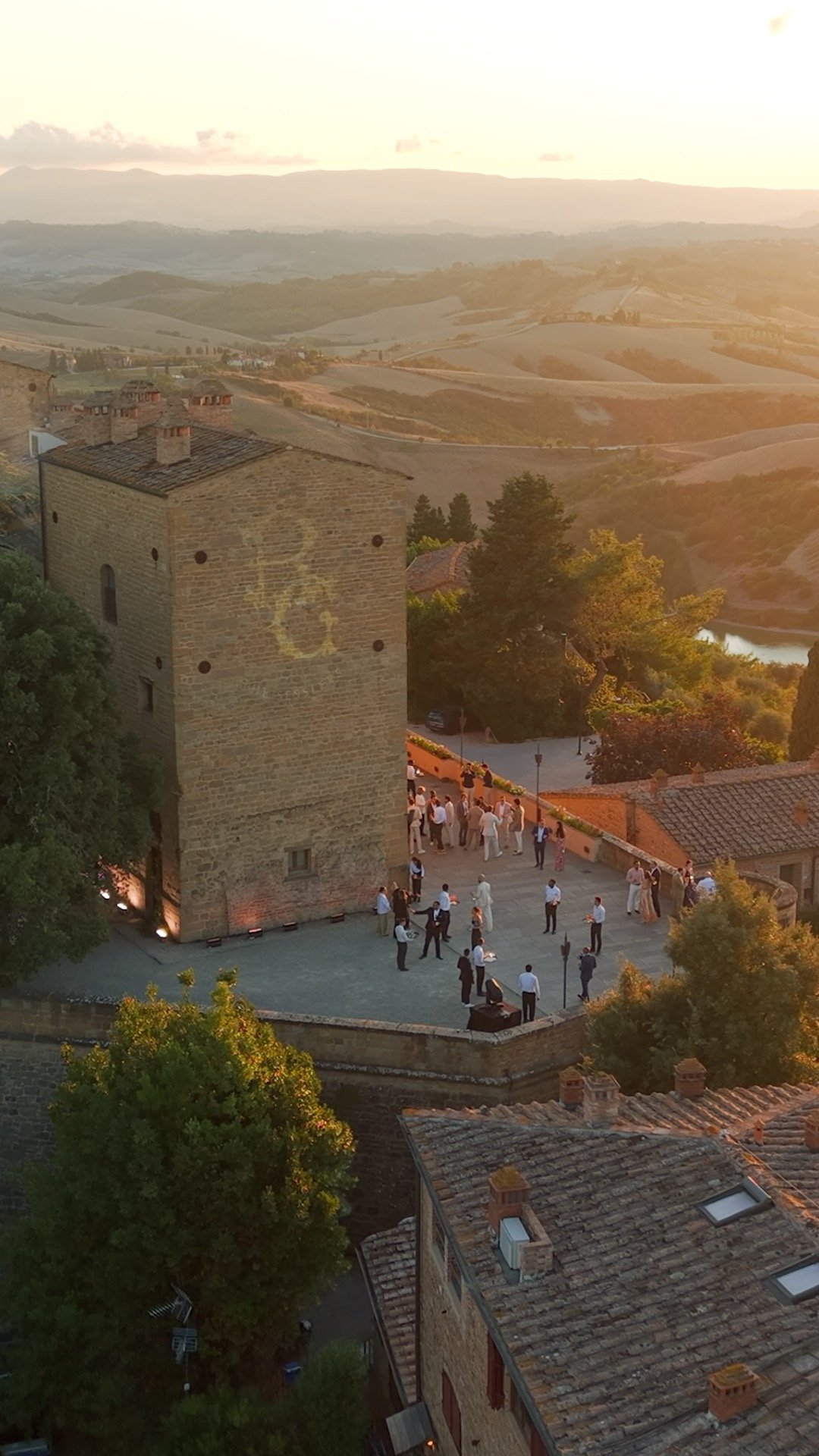 A group of people dressed in formal attire gathering outside near a historic stone tower at sunset, with rolling hills and a lake in the background.