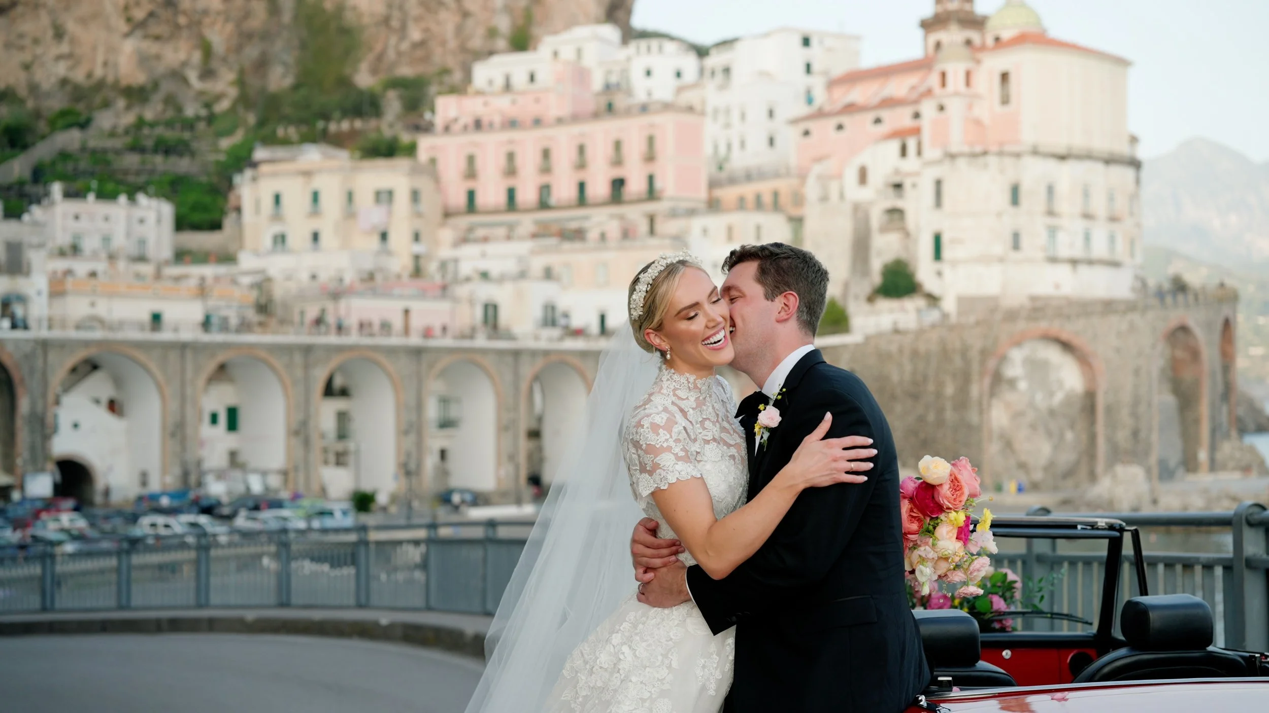 A bride and groom embrace and smile during their wedding photo shoot in front of a colorful hillside town with pastel buildings and an arched bridge.