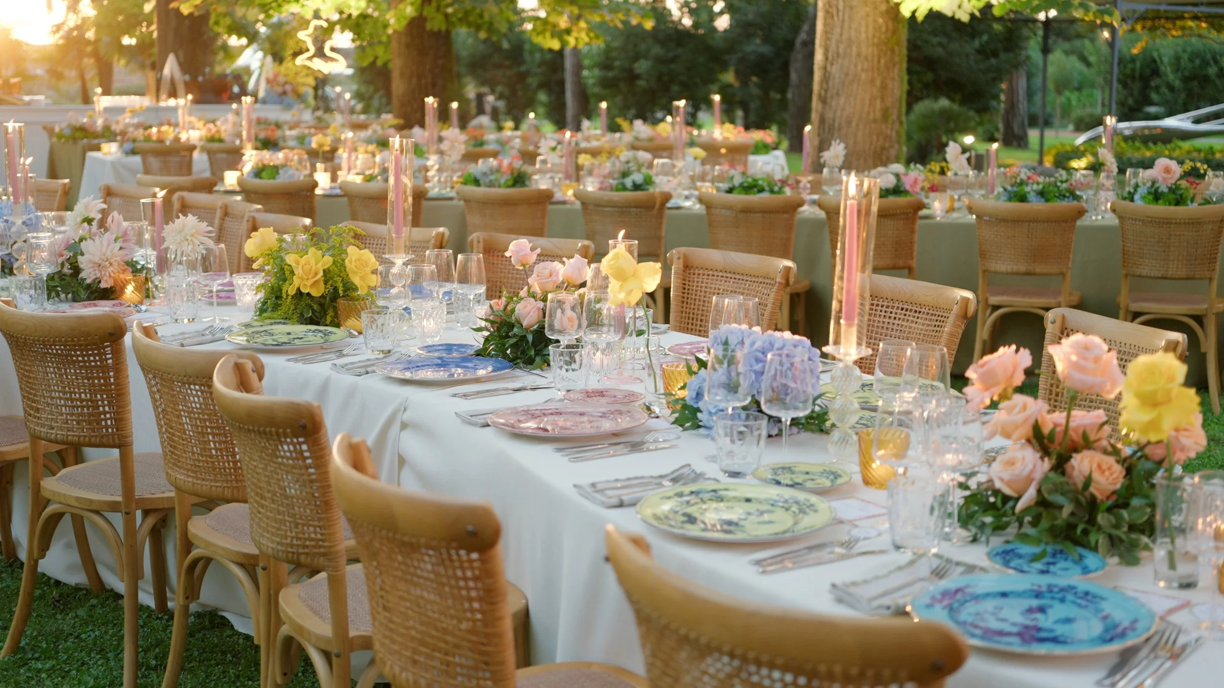 Elegant outdoor dining setup with long table, floral centerpieces, fine china, glassware, and candles, surrounded by wicker chairs, in a lush green garden with trees in the background, during sunset.