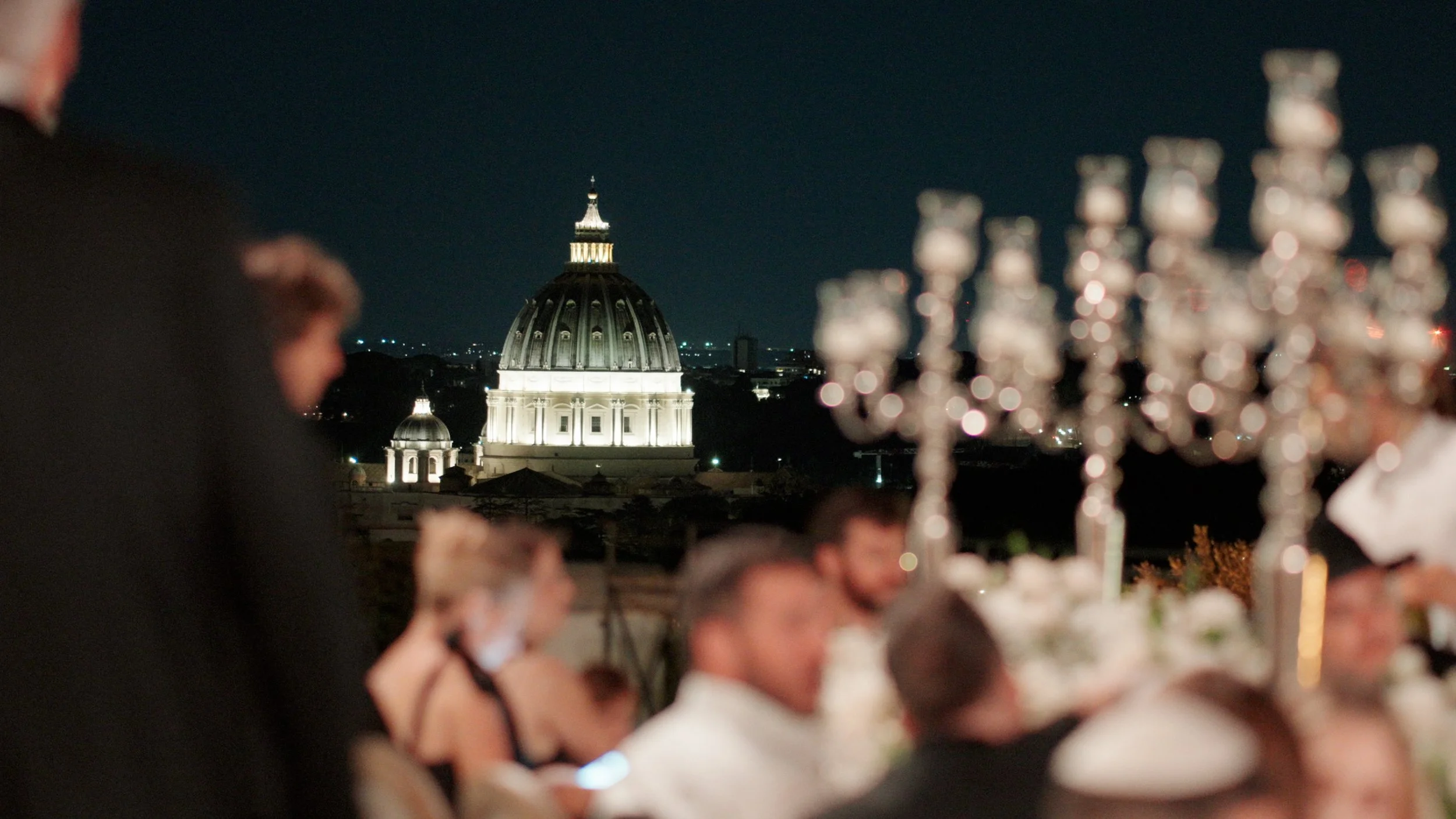 Nighttime scene of a wedding reception outdoors with guests seated at a table with candlesticks and a chandelier, and Saint Peter's Basilica illuminated in the background.