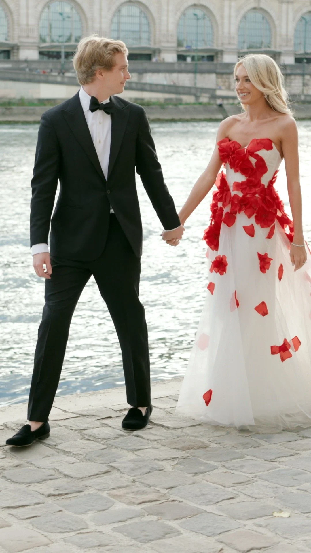 A couple dressed in formal attire, walking hand in hand along a riverside, with a cityscape background.