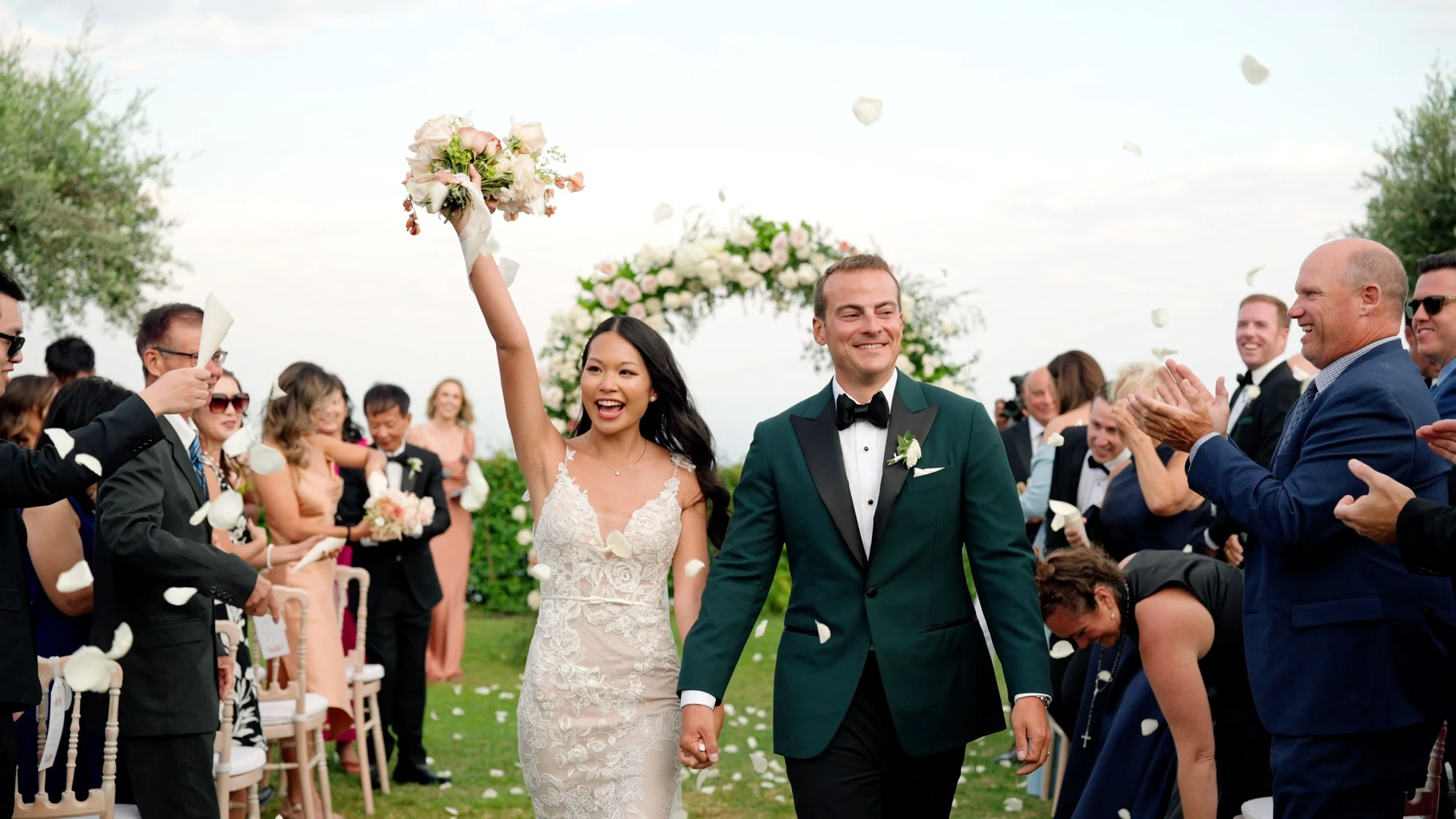 A newlywed couple walking hand-in-hand down the aisle, smiling, as guests celebrate and throw flower petals at an outdoor wedding ceremony.