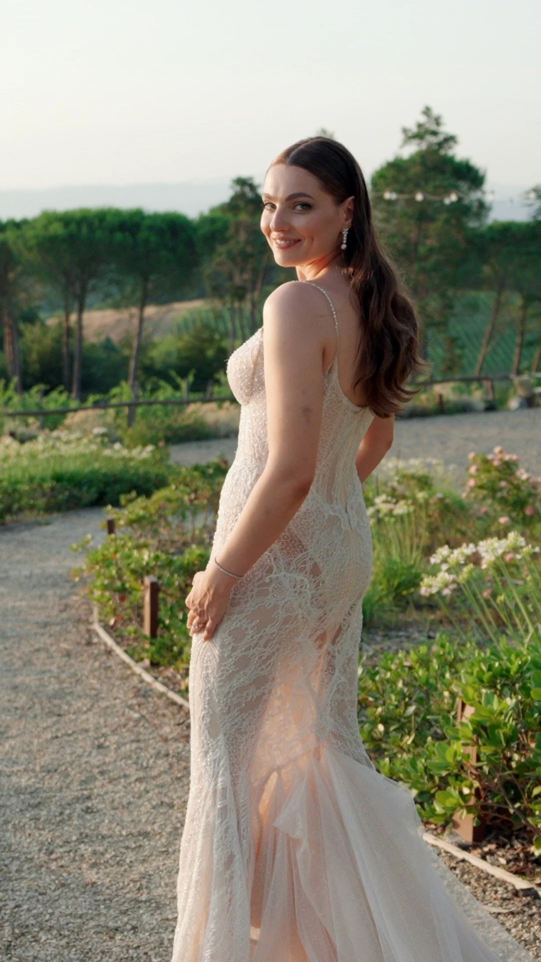 A woman in a white lace wedding dress standing outdoors on a garden path, smiling at the camera.