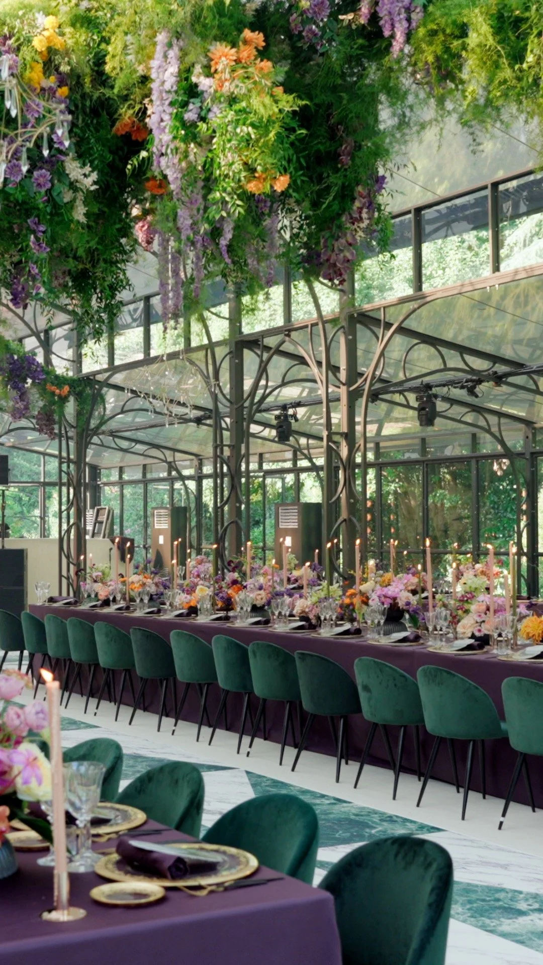 Elegant indoor event with a long table decorated with purple tablecloth, floral centerpieces, and tall pink candles. Green velvet chairs line the sides, and overhead hanging floral arrangements add a lush touch to the glass-enclosed space with a garden view.