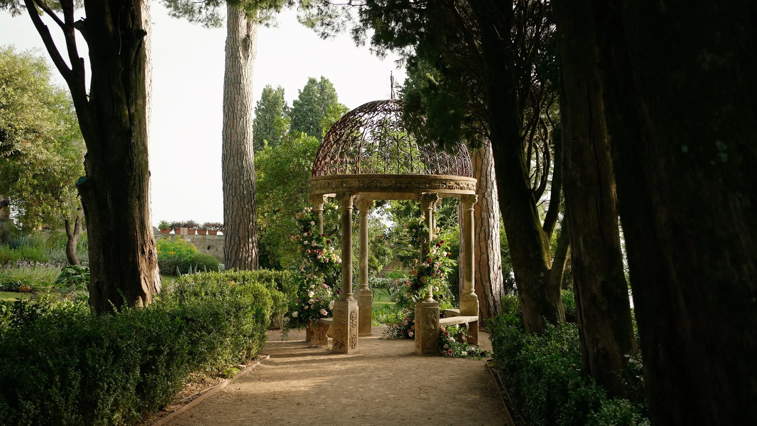 A wedding arch decorated with flowers in a lush, green garden surrounded by tall trees.