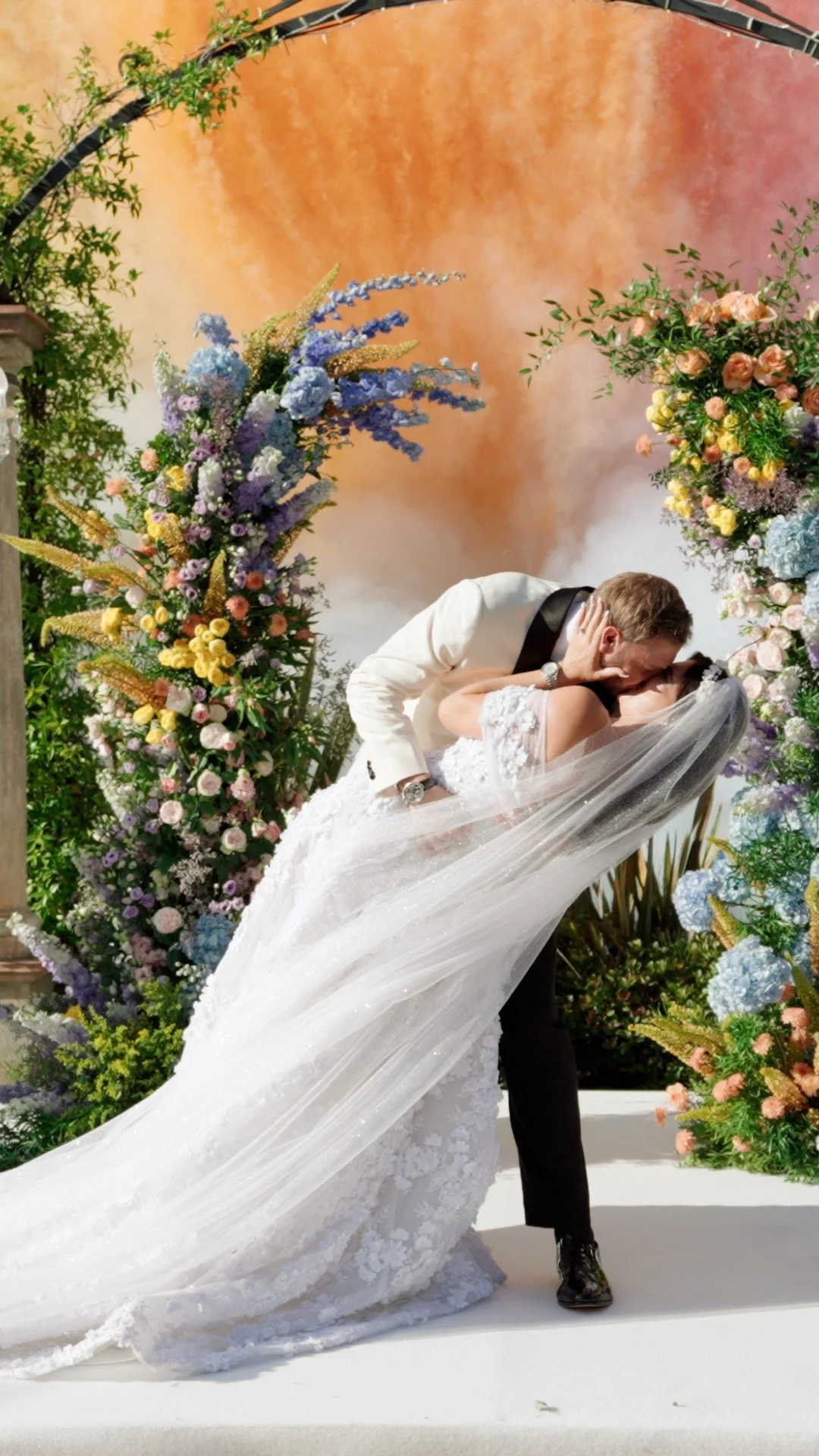 A bride and groom sharing a kiss during their wedding ceremony, surrounded by colorful floral arrangements and a decorative backdrop.