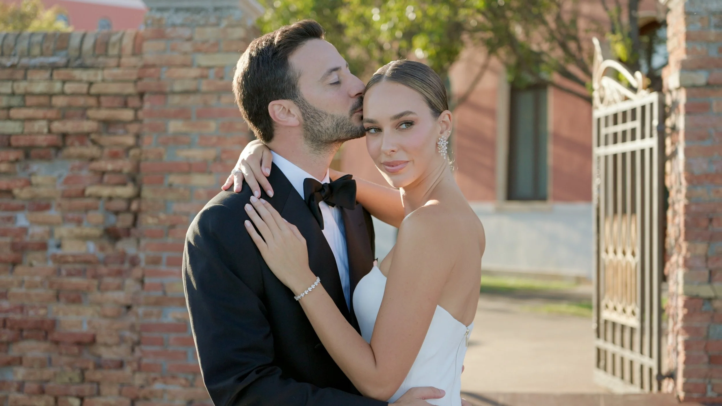 A newlywed couple embracing outdoors, with the groom kissing the bride's temple and the bride looking at the camera, both dressed in wedding attire, standing near a brick wall and an iron gate.