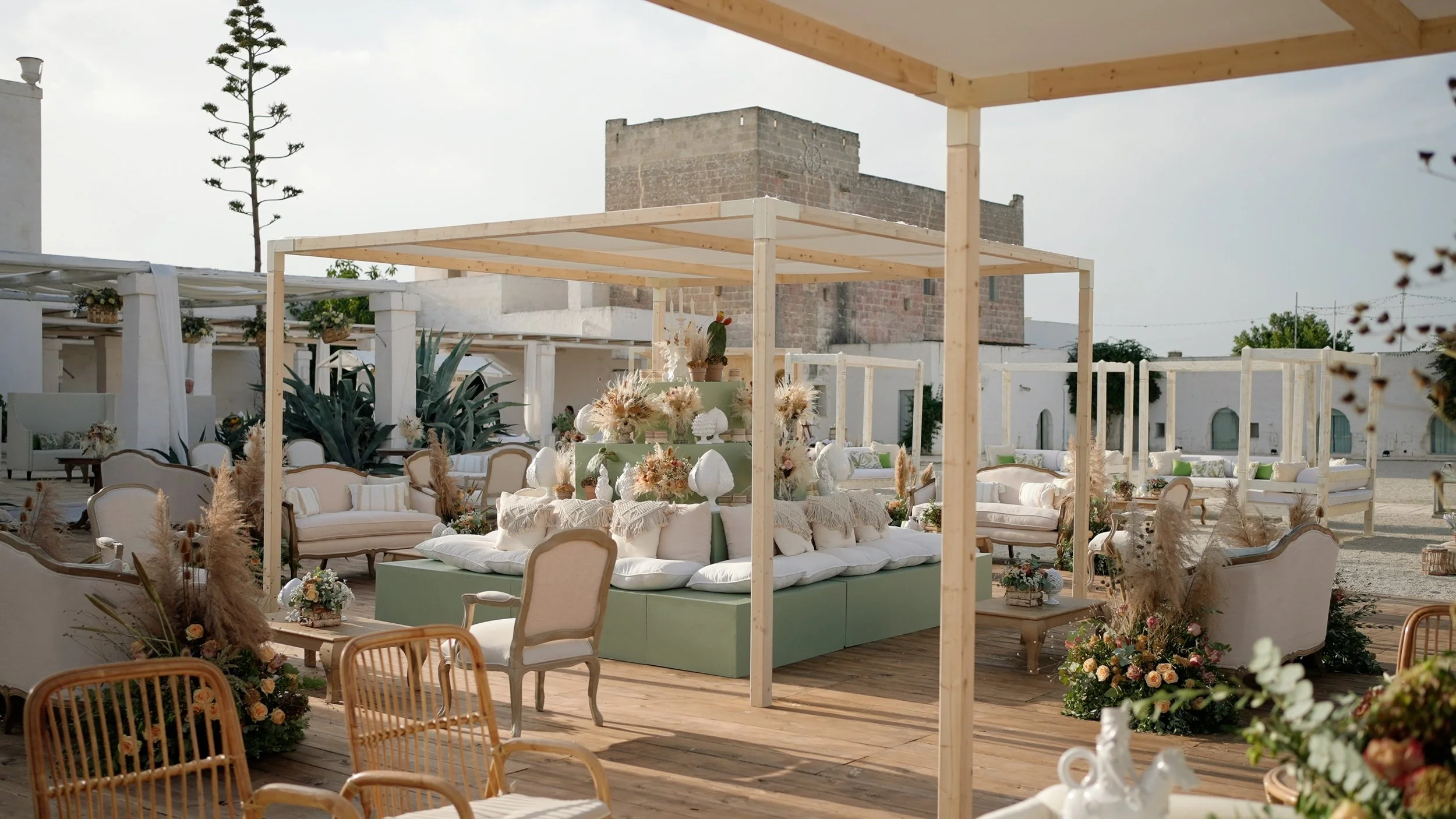 Outdoor event space decorated with pastel-colored furniture, floral arrangements, and wooden canopy structures, with a rustic building and plants in the background.
