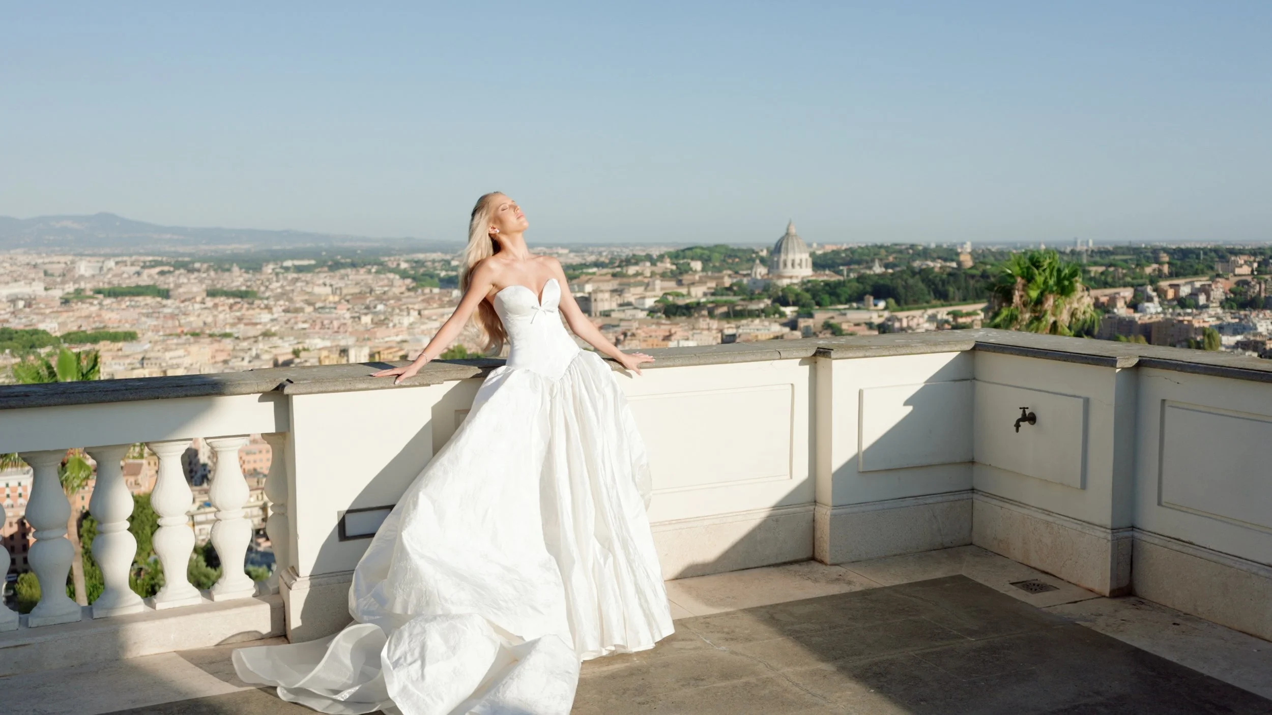 A woman in a white strapless wedding dress with a full skirt stands on a balcony overlooking a cityscape with a domed building in the background. She has her eyes closed and face tilted upward toward the sunlight.