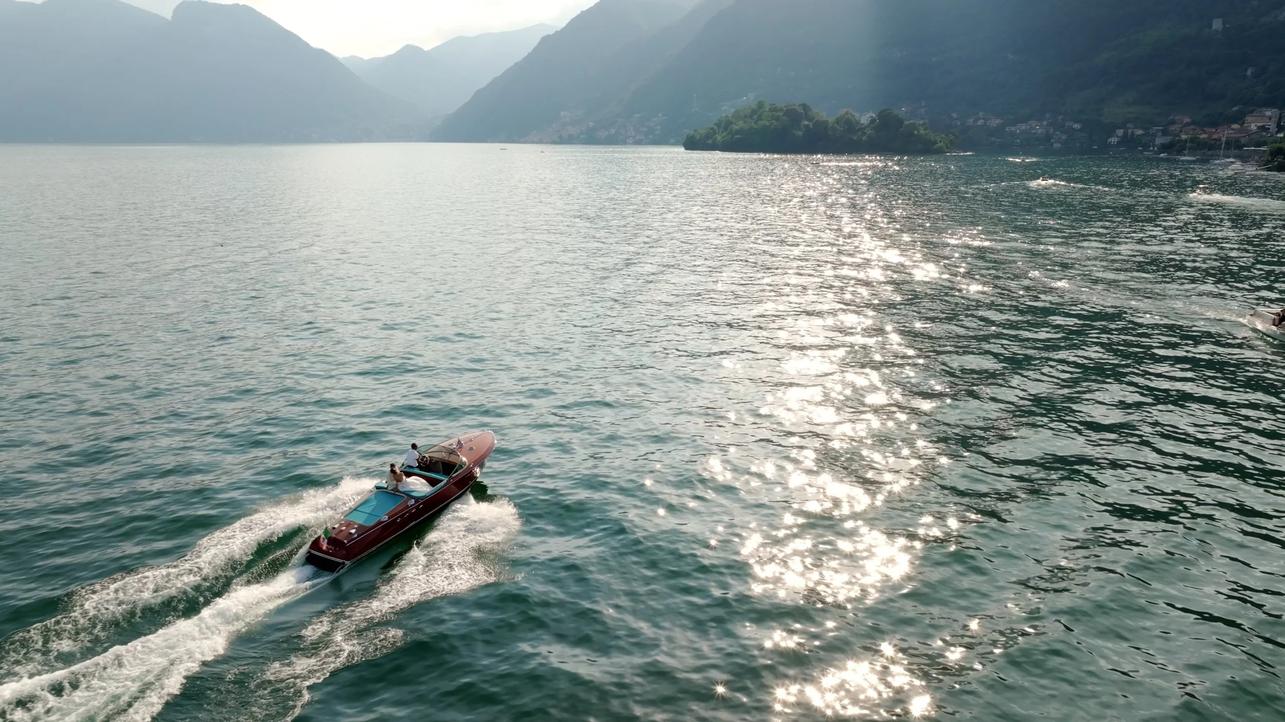 A speedboat with two passengers is traveling across a large lake with mountains in the background; sunlight reflects off the water.