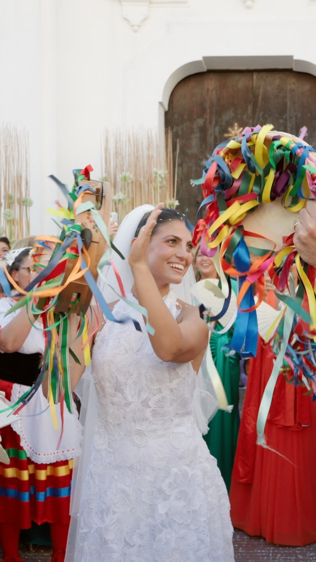 A bride smiling and tossing colorful ribbons during a celebration, with women in traditional dresses around her.