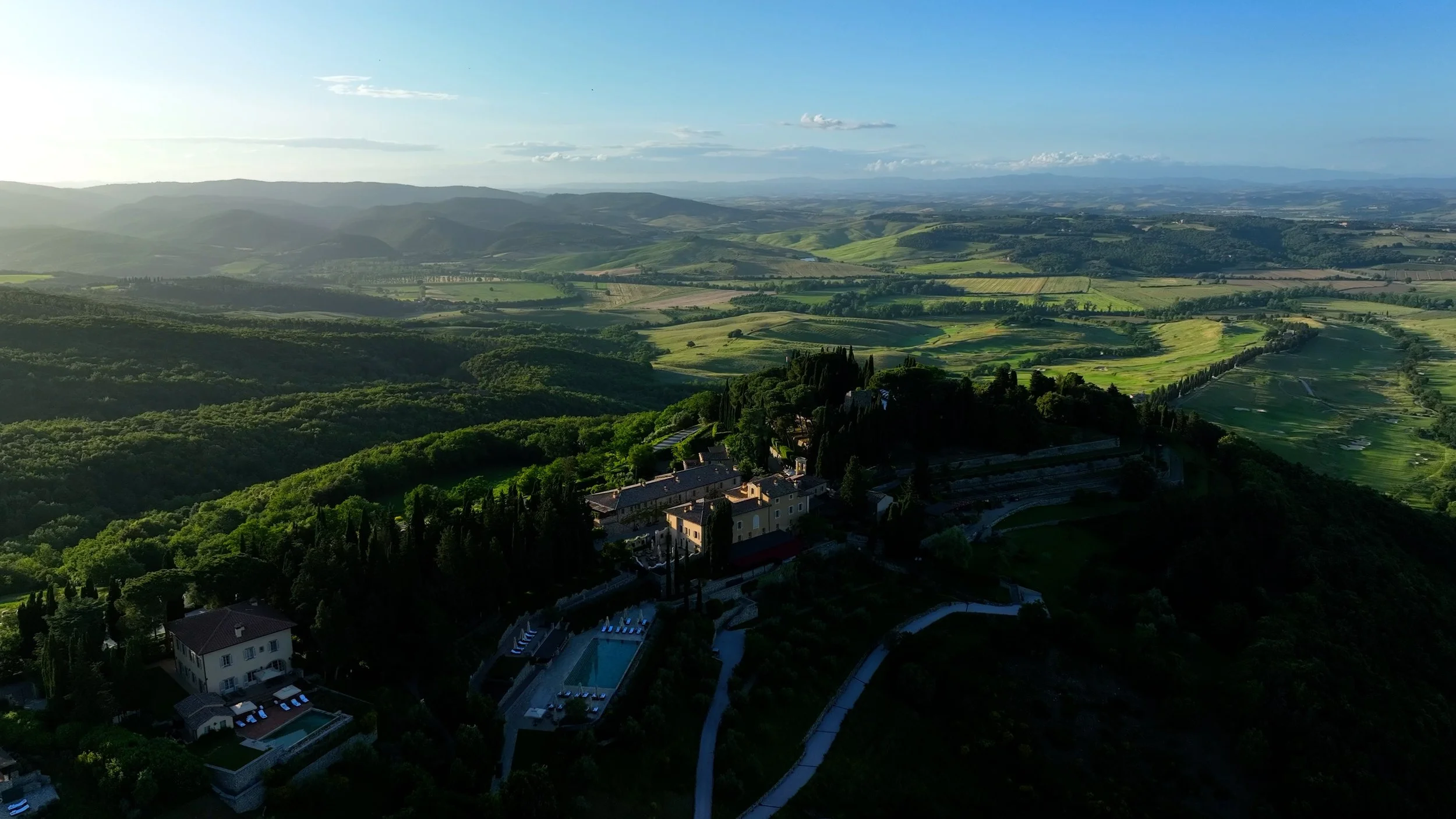 Aerial view of a hilltop estate with buildings, a swimming pool, and surrounding lush green trees, overlooking rolling green hills and farmland under a clear blue sky.
