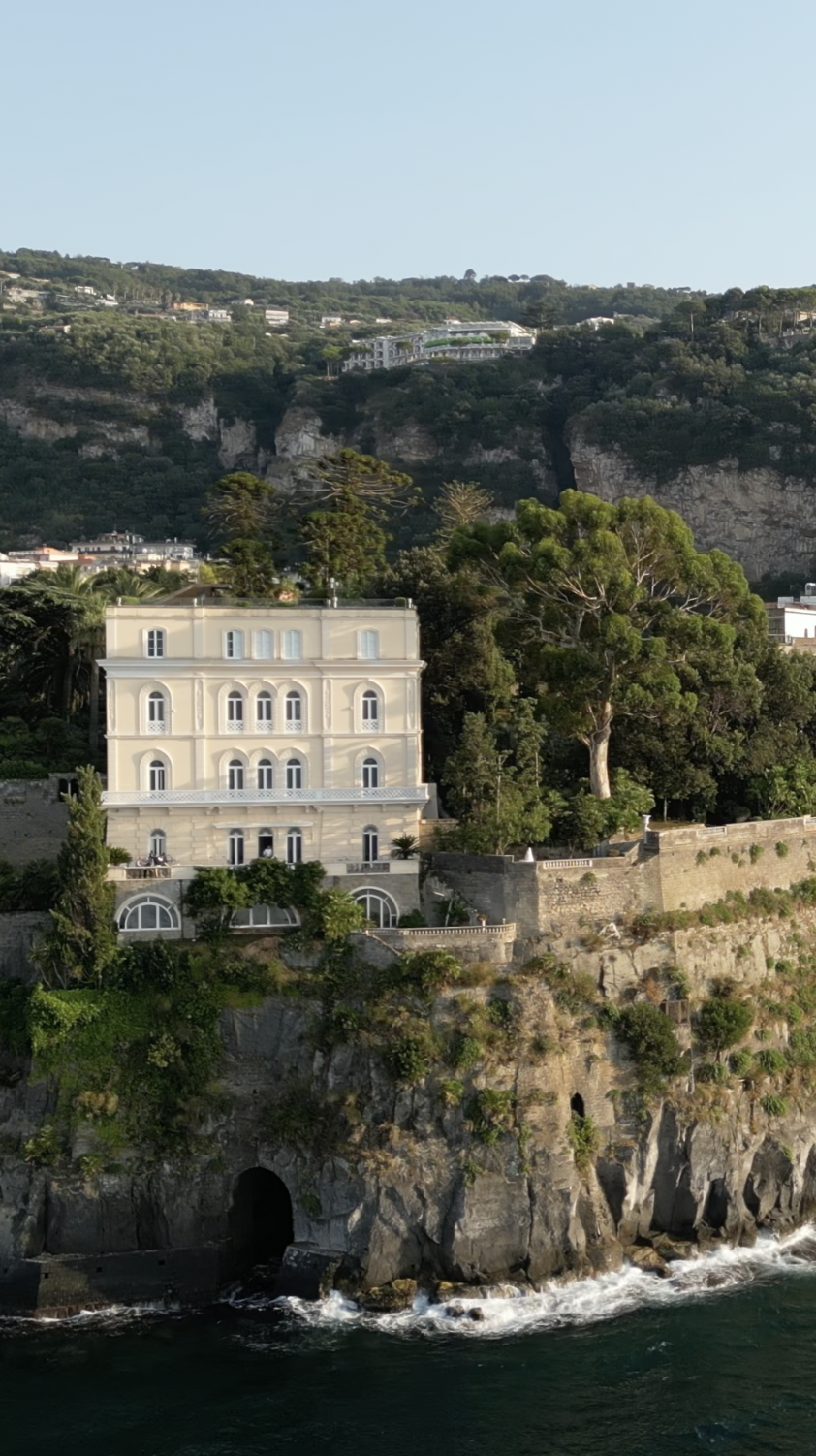 A large, elegant white house built on a rocky cliffside overlooking the ocean, with lush green trees surrounding it and other homes on the hillside in the background.