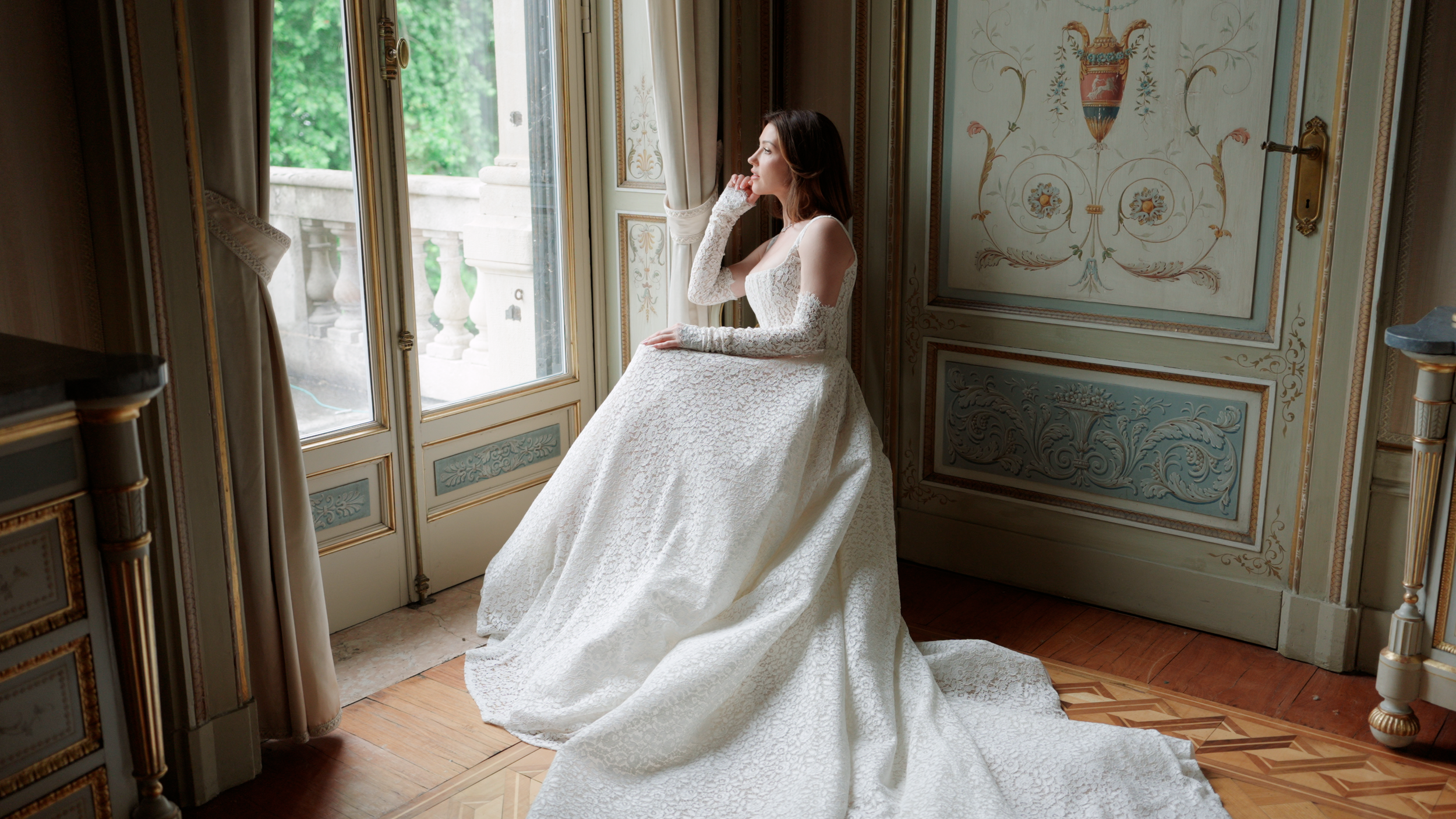A woman in a wedding dress sitting by a large window, looking outside, in an elegant, ornately decorated room.