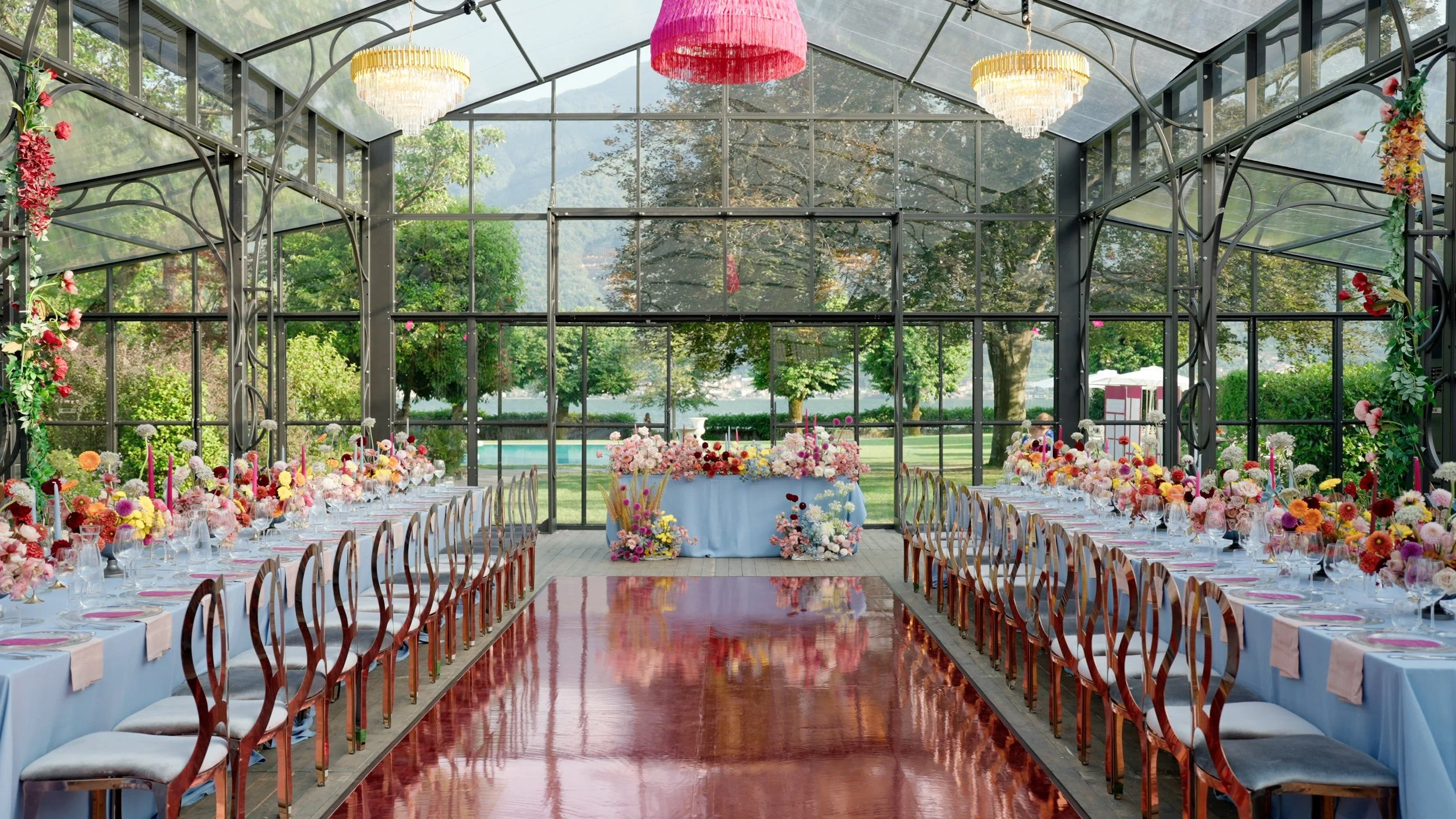 Elegant indoor dining setup with long tables adorned with colorful flower arrangements and candles, enclosed in a glass greenhouse with outdoor greenery visible through the windows.