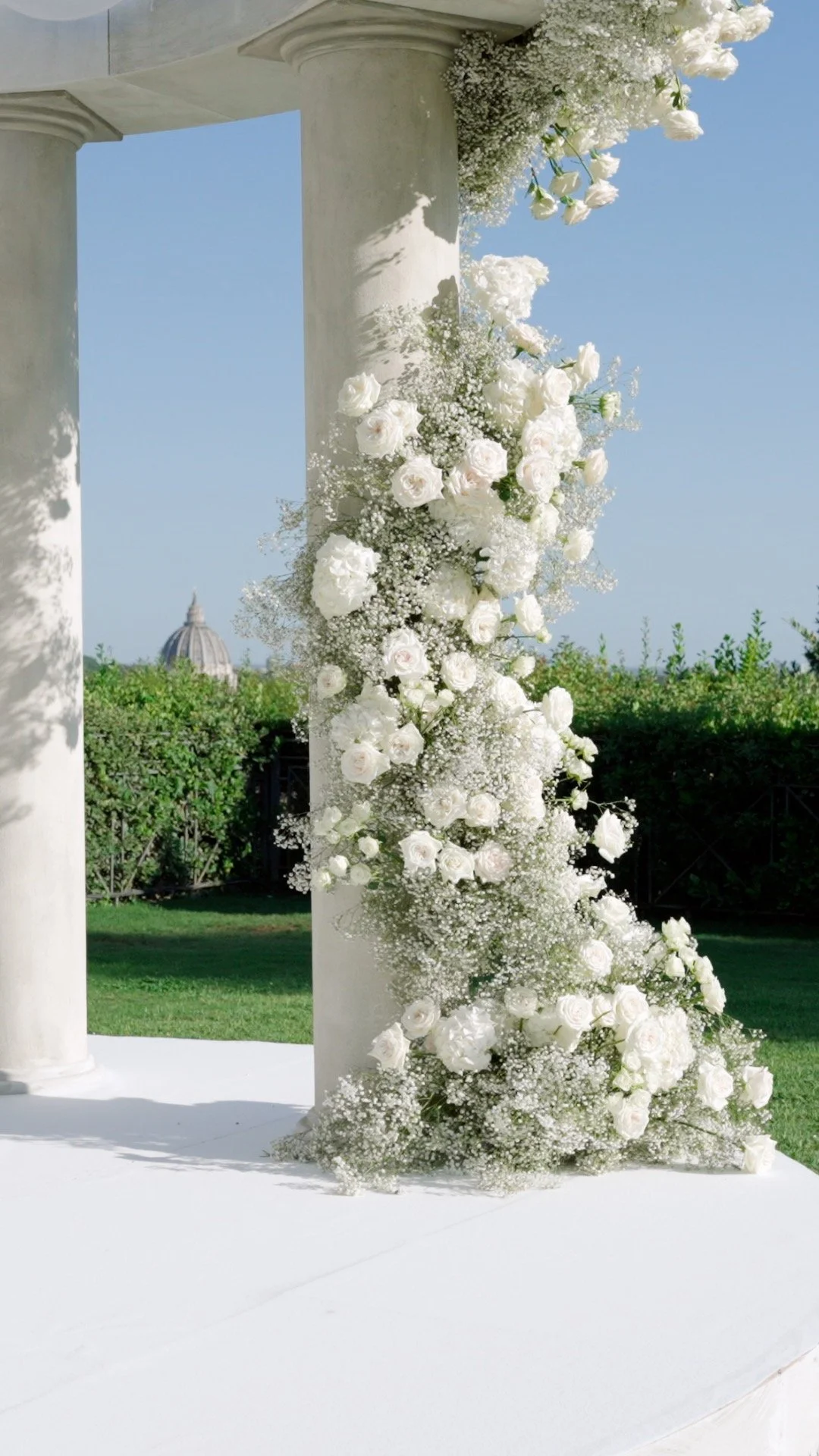 Wedding arch decorated with white roses and baby's breath, set outdoors with a clear blue sky and greenery in the background.