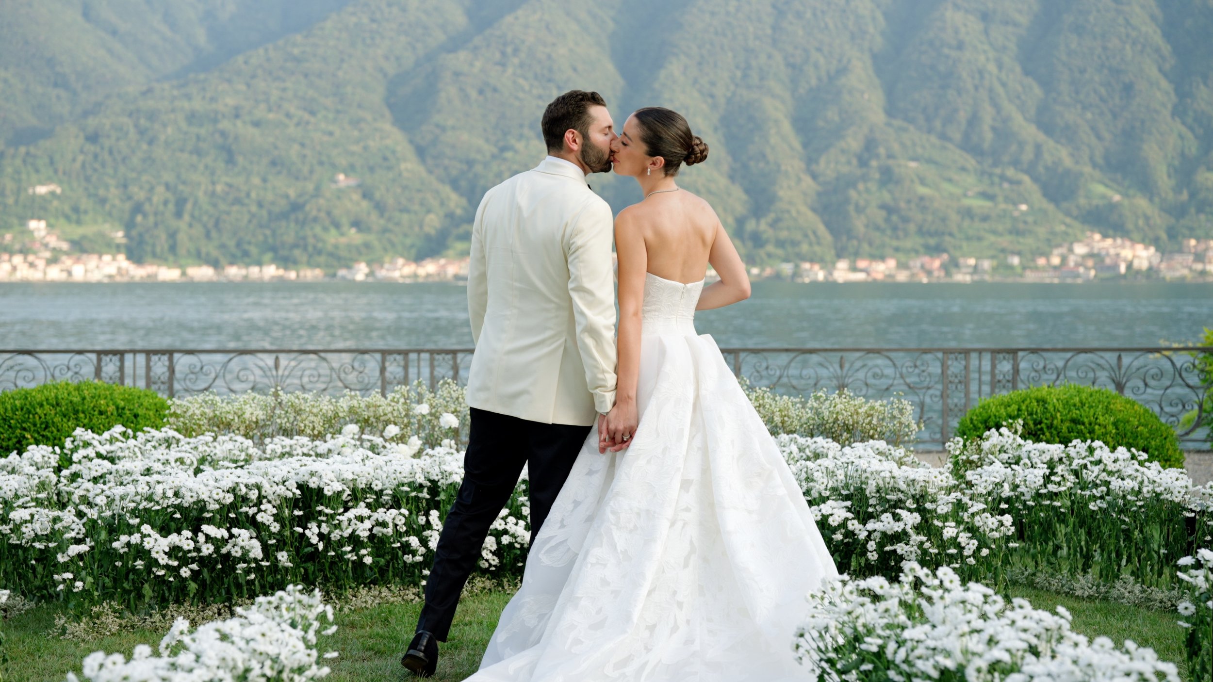 A newlywed couple is kissing in a garden by a lakeside with snowy mountains in the background. The bride is in a white strapless wedding gown, and the groom is in a white suit jacket and dark pants. White flowers and green bushes surround them.