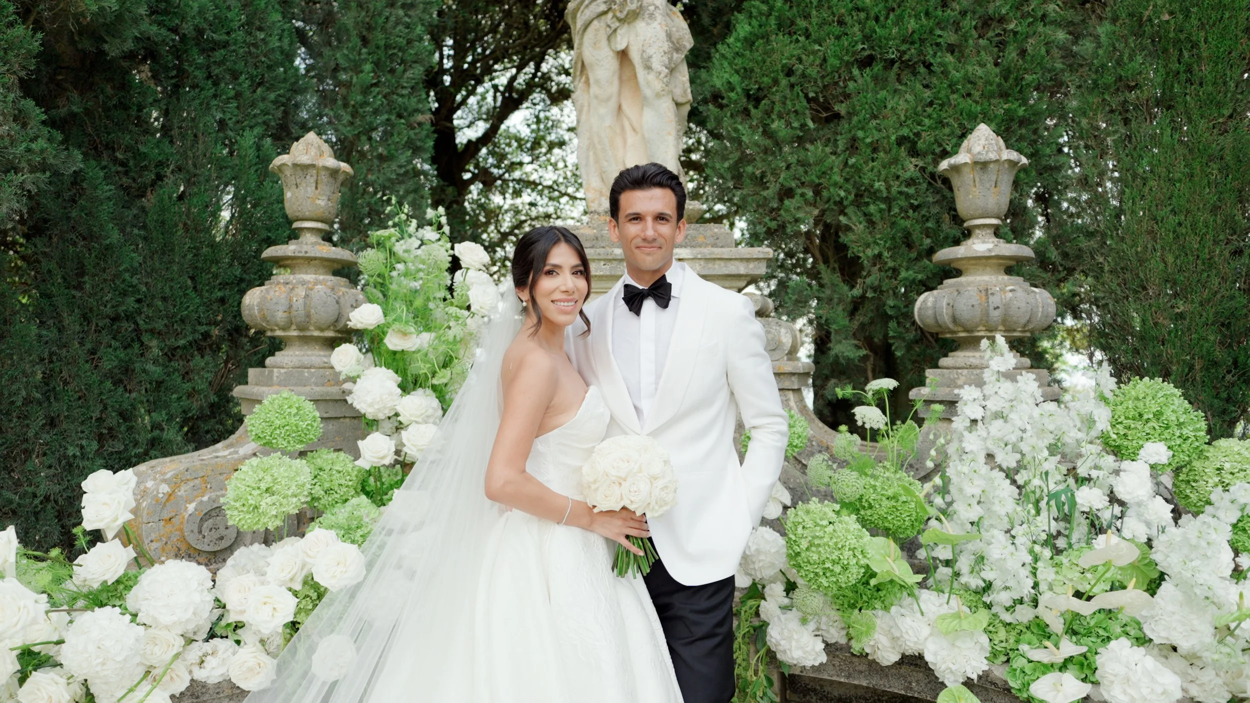 A bride and groom standing outdoors in front of a statue and surrounded by white and green flowers, dressed in wedding attire, smiling at the camera.