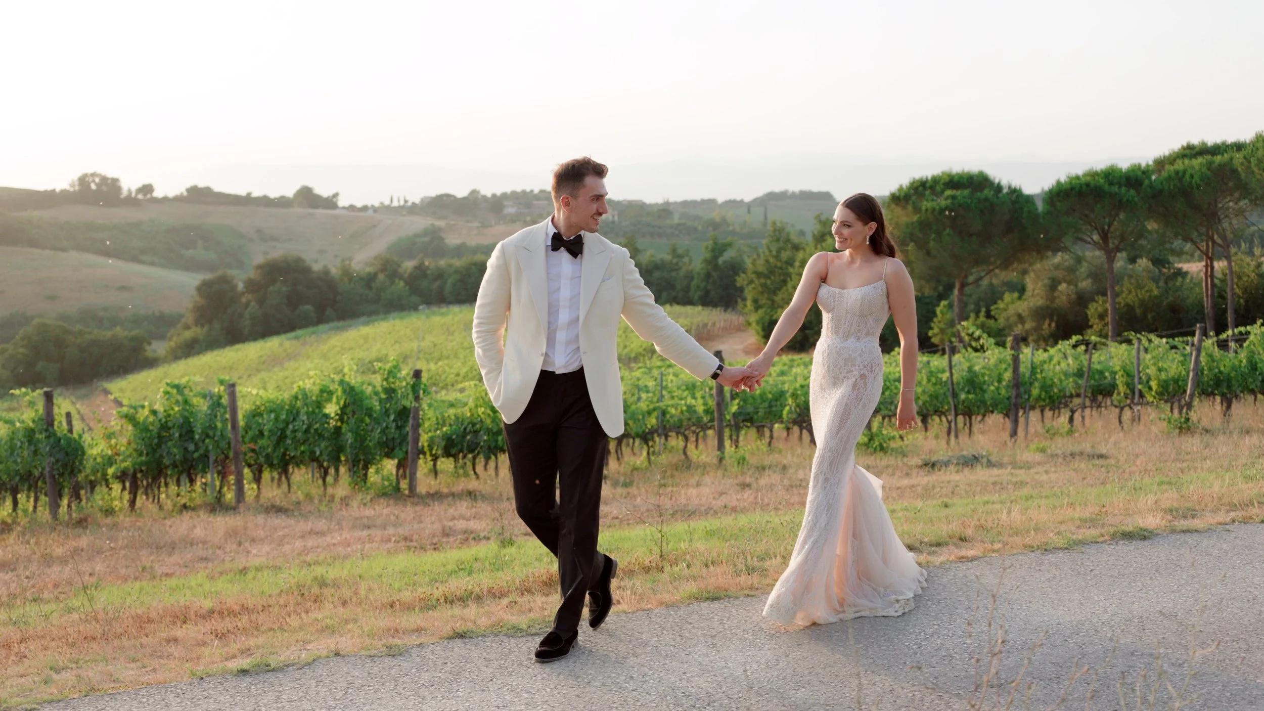 A newlywed couple walking hand in hand along a vineyard path during sunset, with green vine-covered hills and trees in the background.