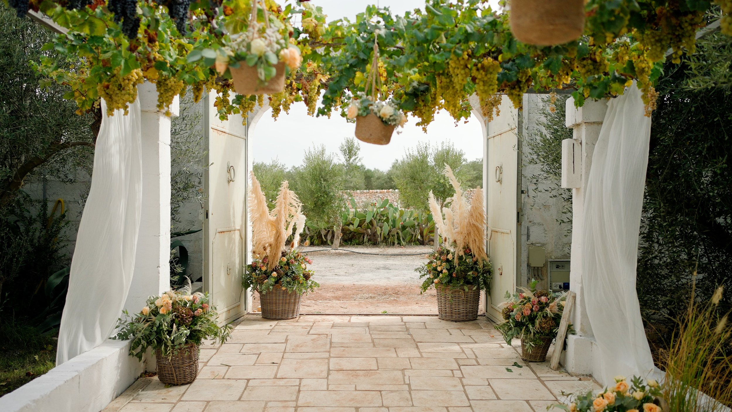 Decorated outdoor gate with floral arrangements, hanging baskets, white curtains, and a garden view with cactus and trees.