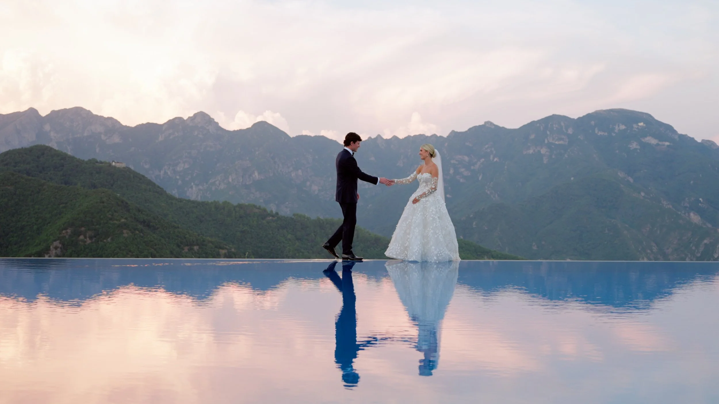 A bride and groom holding hands on an infinity pool overlooking mountainous landscapes at sunset.