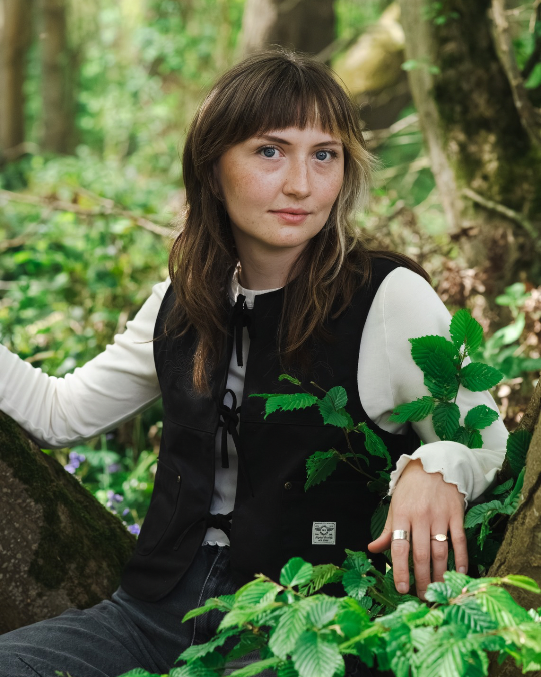 A young woman with brown hair and blue eyes sitting outdoors among green trees and foliage, wearing a black vest over a white long-sleeve shirt, looking slightly to the side.