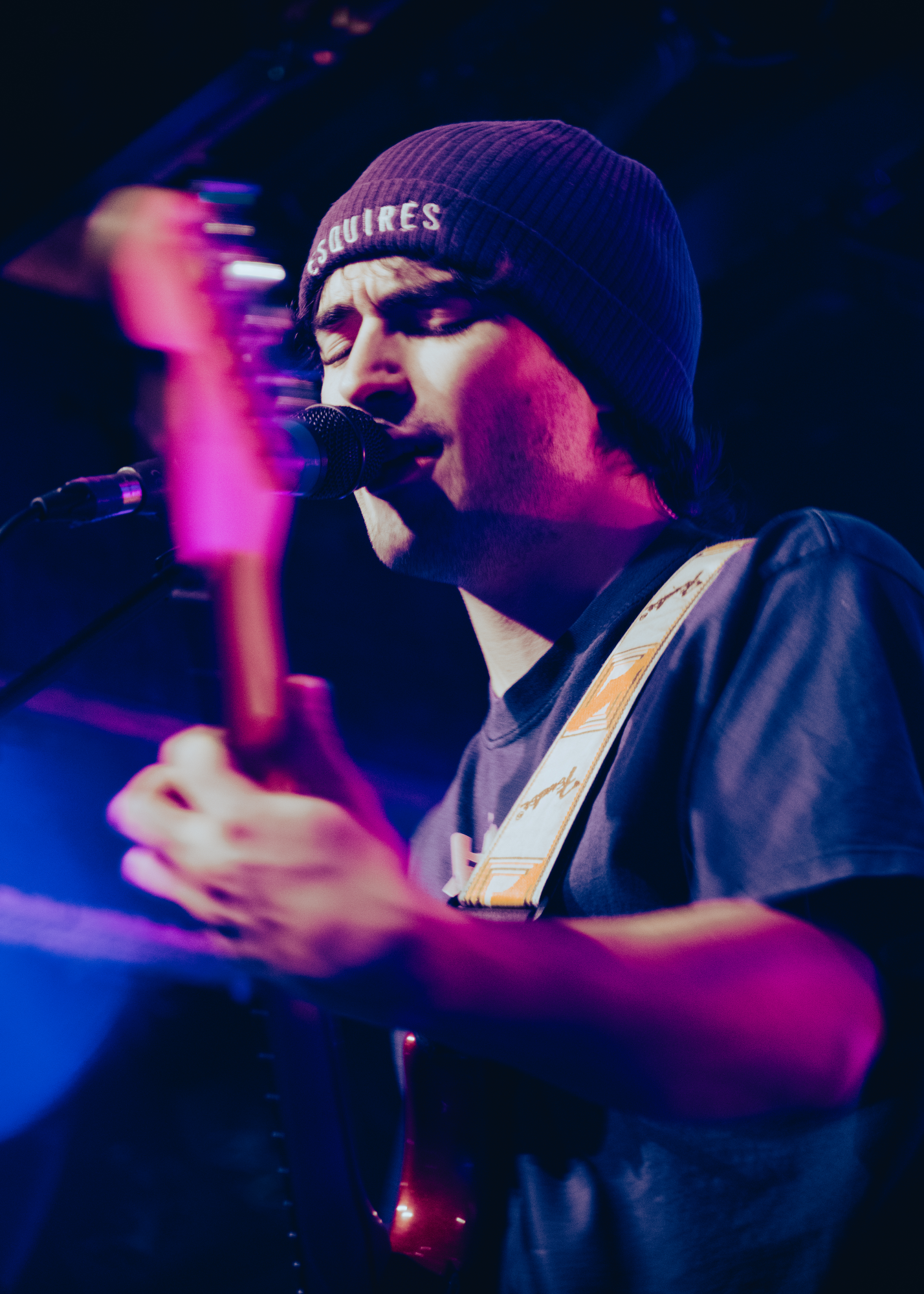 A male musician wearing a beanie and a dark shirt singing into a microphone on stage, with blue and pink stage lighting.